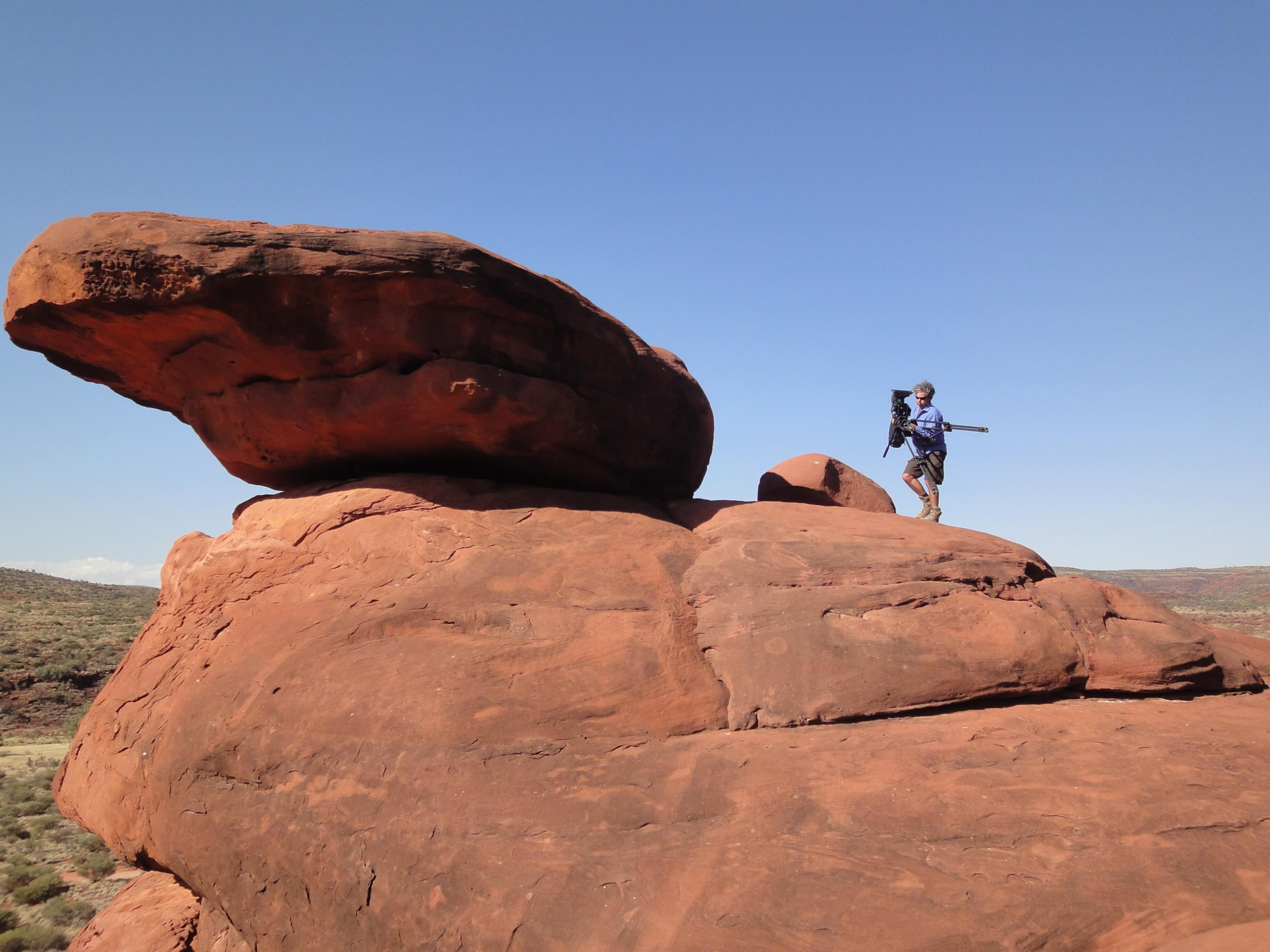 A person stands on a large red rock formation, holding a hiking pole and gazing out at the Terra Mater landscape. The clear blue sky enhances the natural beauty of the scene, offering profound insights into the terrain below—a harmonious blend of desert shrubs and rocks.