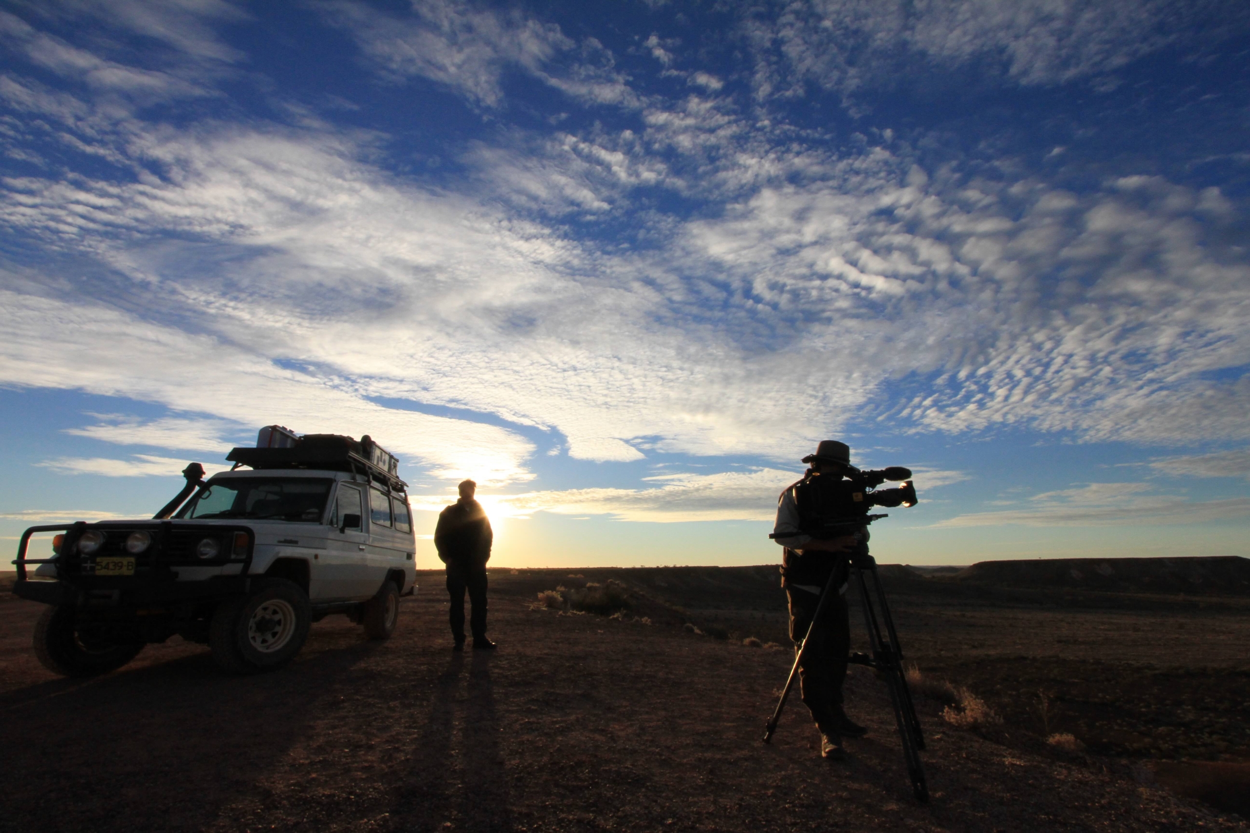 Two people with a white SUV set up a camera tripod on a desert road at sunset, capturing the breathtaking beauty of Terra Mater under a vast, cloud-filled sky.