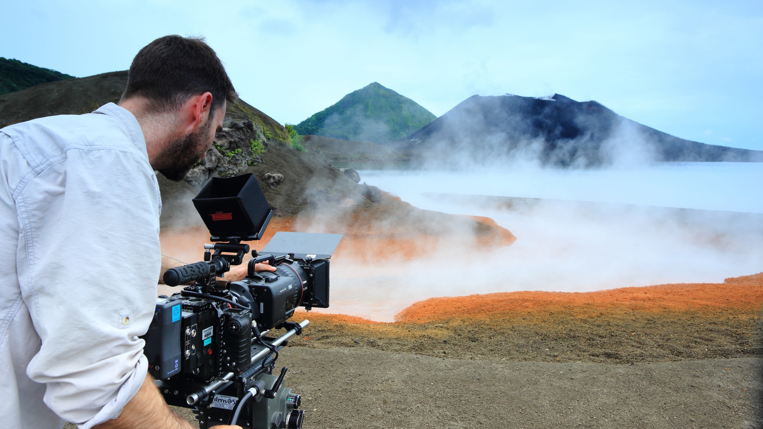 A person operates a video camera on a tripod overlooking the volcanic landscape, capturing insights inspired by Terra Mater. Steam rises from the multicolored earth near a caldera, with hills visible in the background under a cloudy sky.