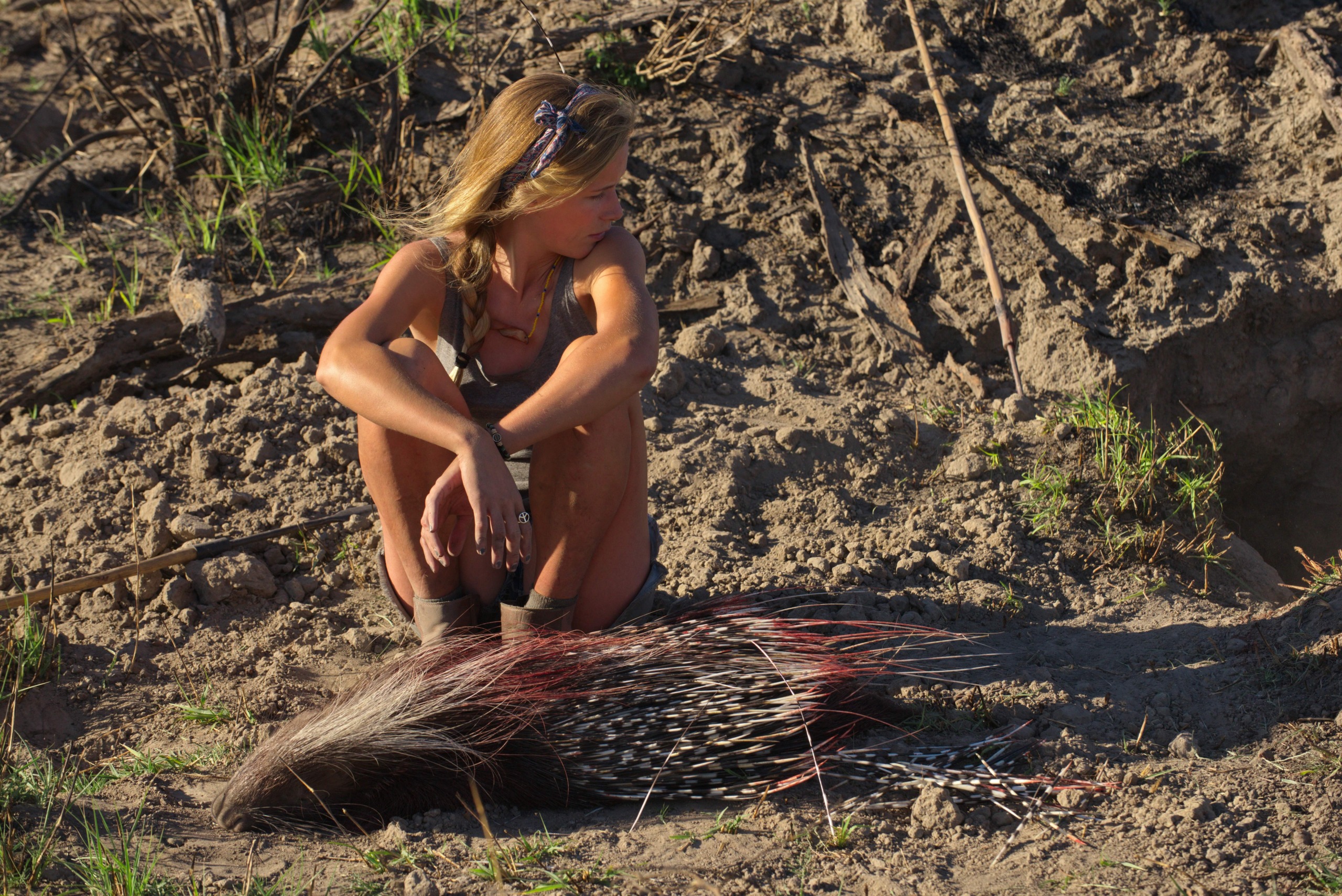 A blonde-haired model sits on the ground in a dry, grassy area, looking to her right. Dressed in shorts and boots, she embodies a modern-day Bushmen style. Beside her lies a large porcupine on its side, enhancing the natural outdoor setting.