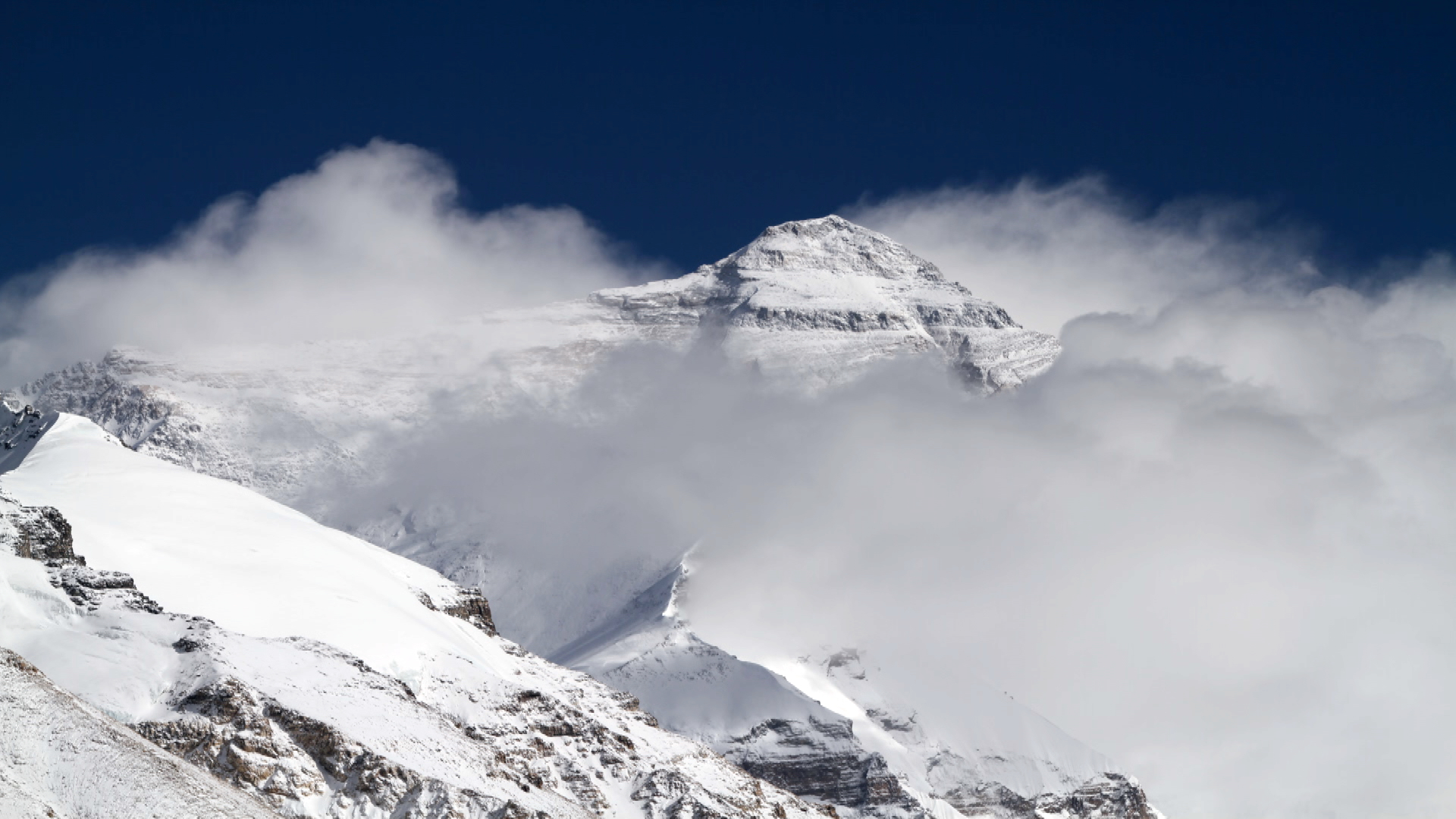 A snow-covered mountain peak, partially wrapped in clouds under a clear blue sky, rises majestically, reminiscent of vistas guided by expert Sherpas.