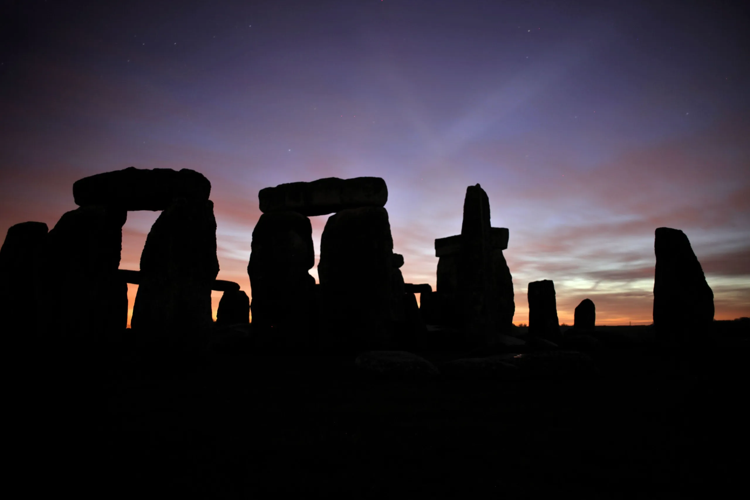 Against a colorful sky transitioning from deep blue to orange and pink hues, the silhouette of Stonehenge stands majestic at twilight. The ancient stone structures appear dark against the vibrant, dusk-lit sky, capturing the site's timeless allure.