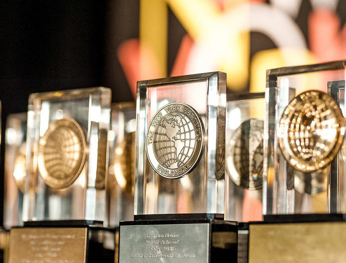 A close-up of several crystal awards with gold medallions inside, arranged in rows. The medallions, reminiscent of the prestigious New York Festivals, feature an embossed globe design. The background is a blurred mix of black and yellow hues, casting a spotlight on the honored productions awarded.