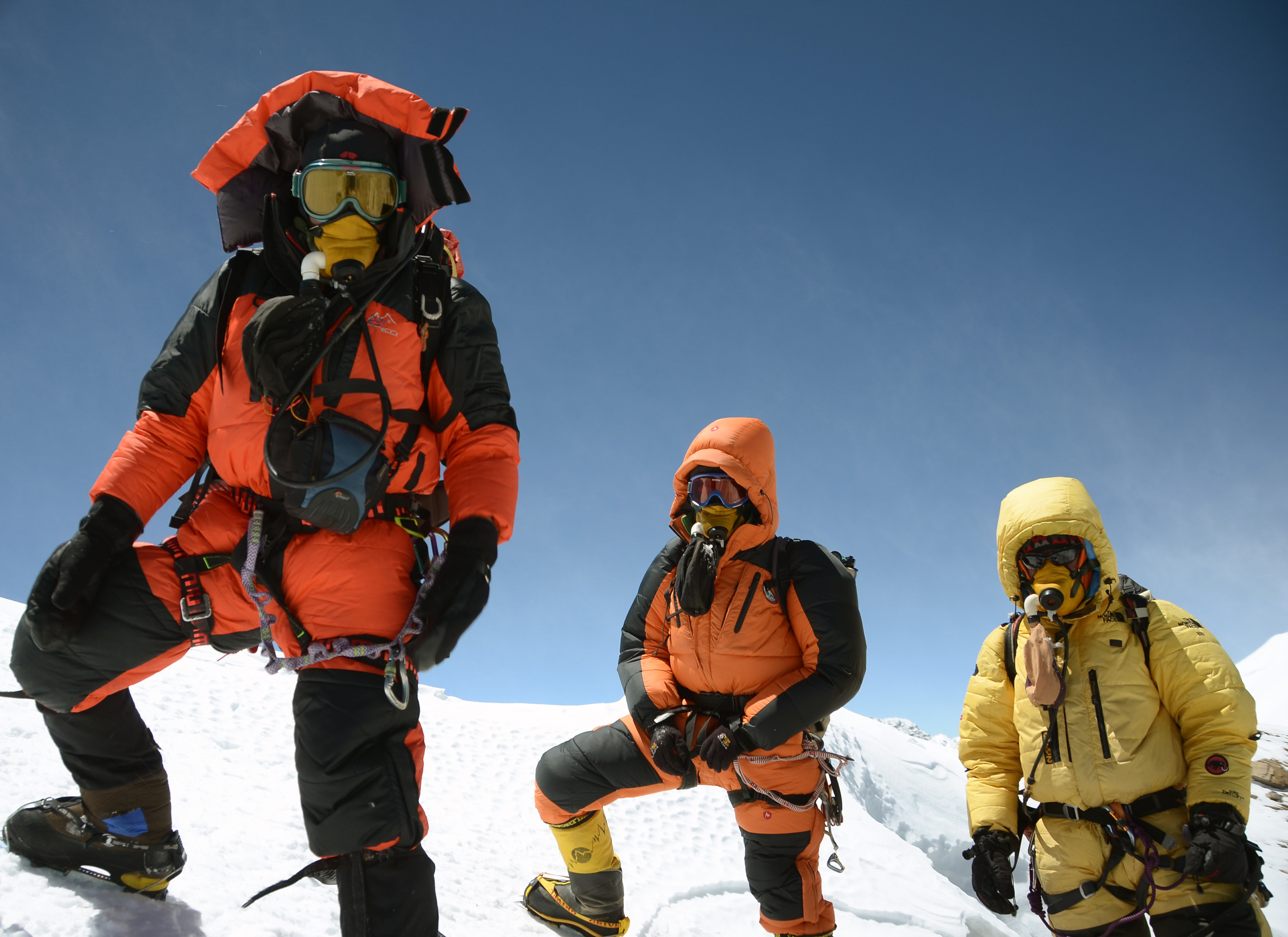 Three mountaineers in colorful climbing suits and oxygen masks stand alongside a skilled Sherpa on a snowy peak against a clear blue sky. They wear helmets and harnesses, indicating high-altitude conditions.