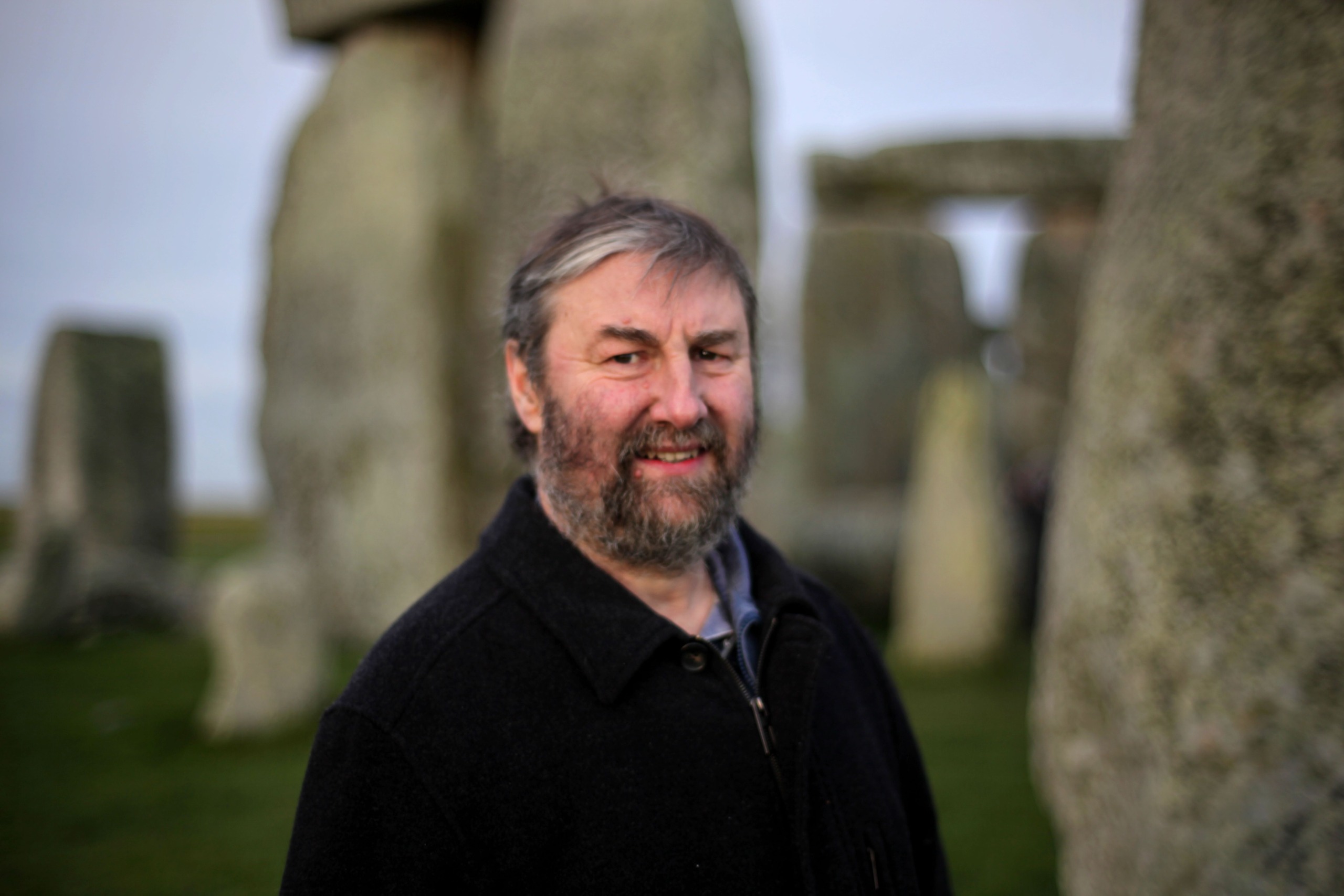 A person with a beard and wearing a dark coat stands outdoors in front of large stone structures, strikingly reminiscent of Stonehenge. The sky is overcast, adding a mystic vibe as the individual smiles warmly at the camera.