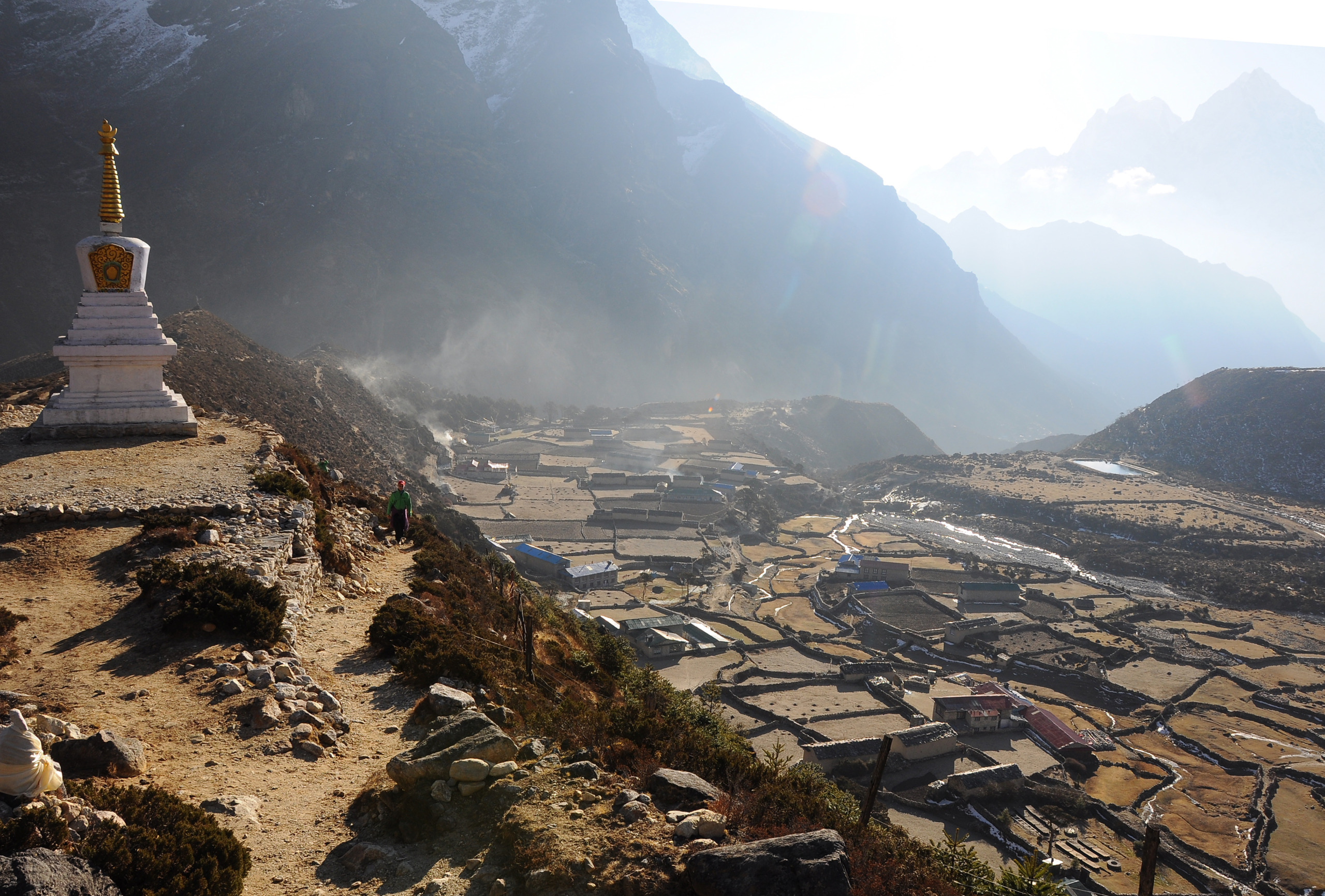 A small, Buddhist stupa stands on a rocky mountain path, often traveled by sherpas, overlooking a valley with clustered buildings. Snow-capped peaks rise in the distance under a hazy sky, casting light across the landscape.