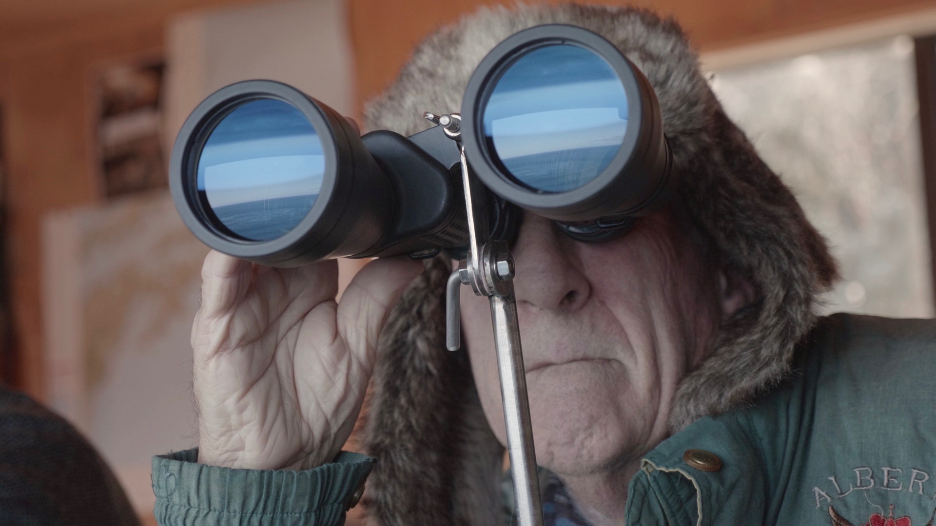 An older person wearing a fur-lined hat and green coat peers through binoculars, searching intently, as if spotting a whale on the distant horizon. Indoors, a map in the background hints at past adventures and future discoveries.