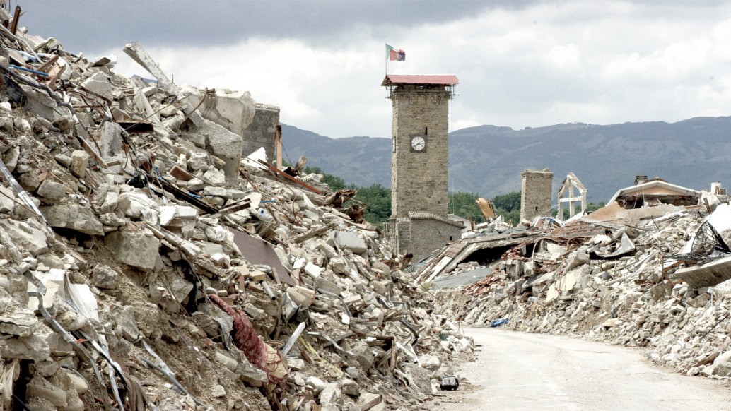 Rubble and debris of collapsed buildings line a street in a red zone village devastated by an earthquake. A clock tower stands amid the destruction under a cloudy sky, with mountains visible in the background, offering a haunting glimpse behind the scenes of this tragedy.