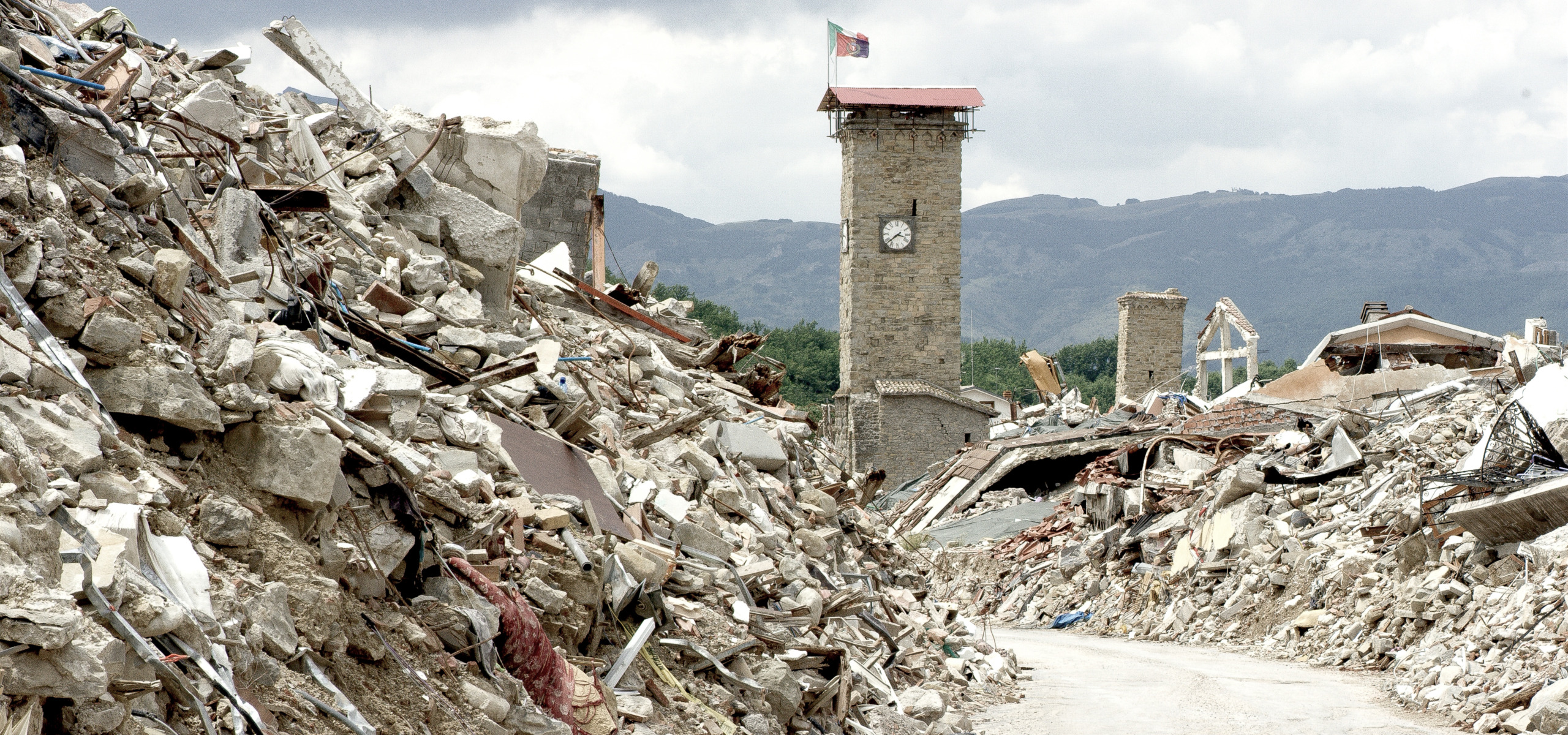 Rubble and debris of collapsed buildings line a street in a red zone village devastated by an earthquake. A clock tower stands amid the destruction under a cloudy sky, with mountains visible in the background, offering a haunting glimpse behind the scenes of this tragedy.