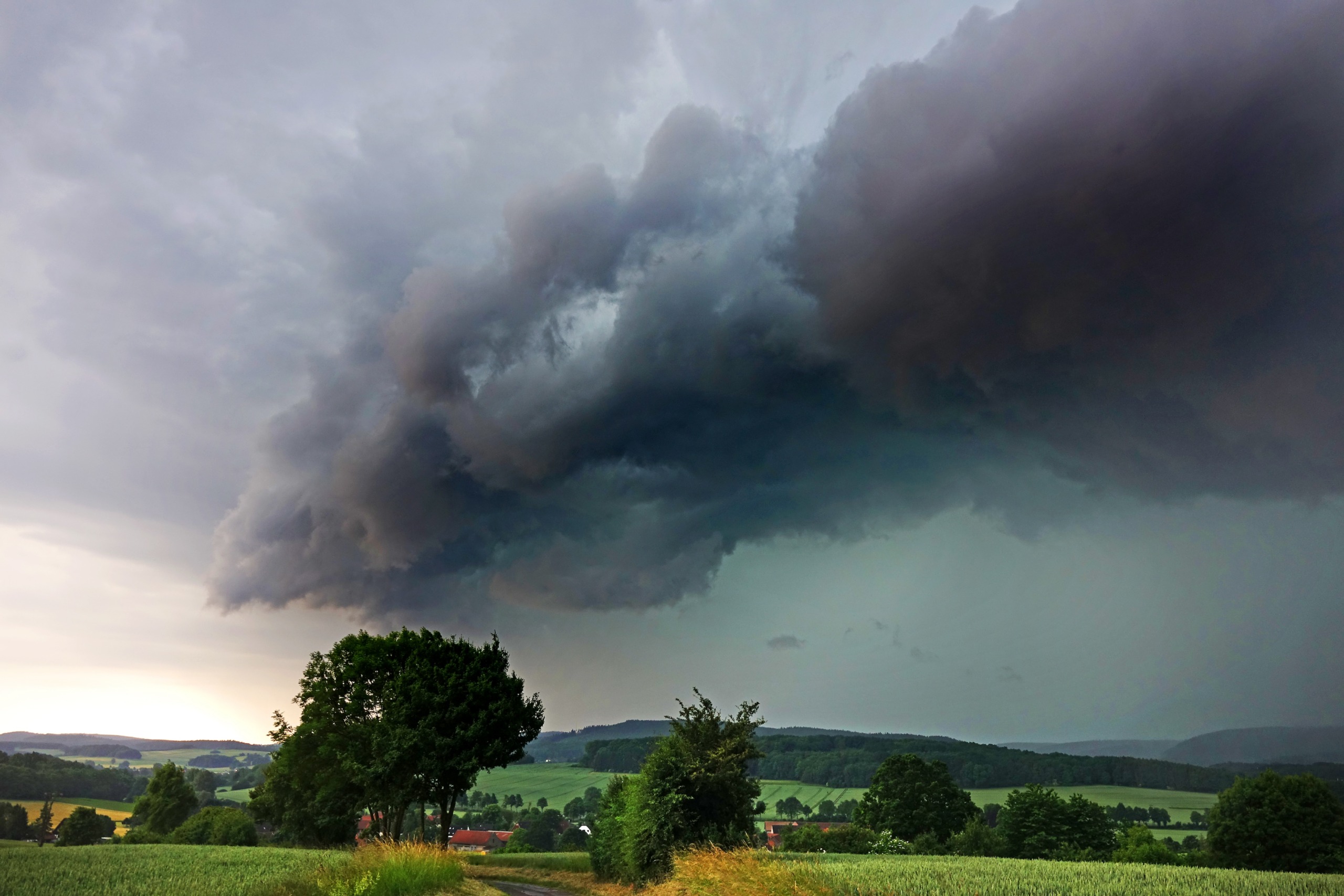 A rural landscape unfolds with a dramatic, dark storm cloud looming over vibrant green fields and trees. The wild weather paints the sky in a mix of dark gray and lighter shades, heralding an impending storm.