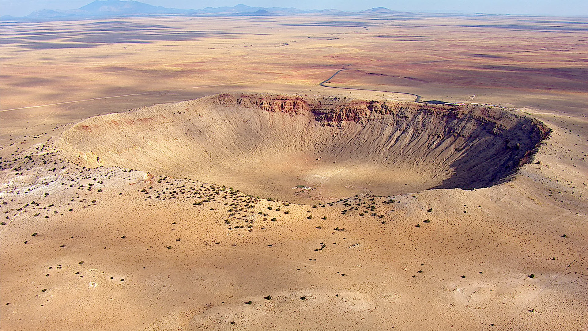 Aerial view of a large meteor crater on Earth in a barren desert landscape, with a circular shape and steep, rocky walls. Sparse vegetation surrounds the crater, and distant mountains are visible on the horizon under a clear sky, showcasing the raw beauty of nature.