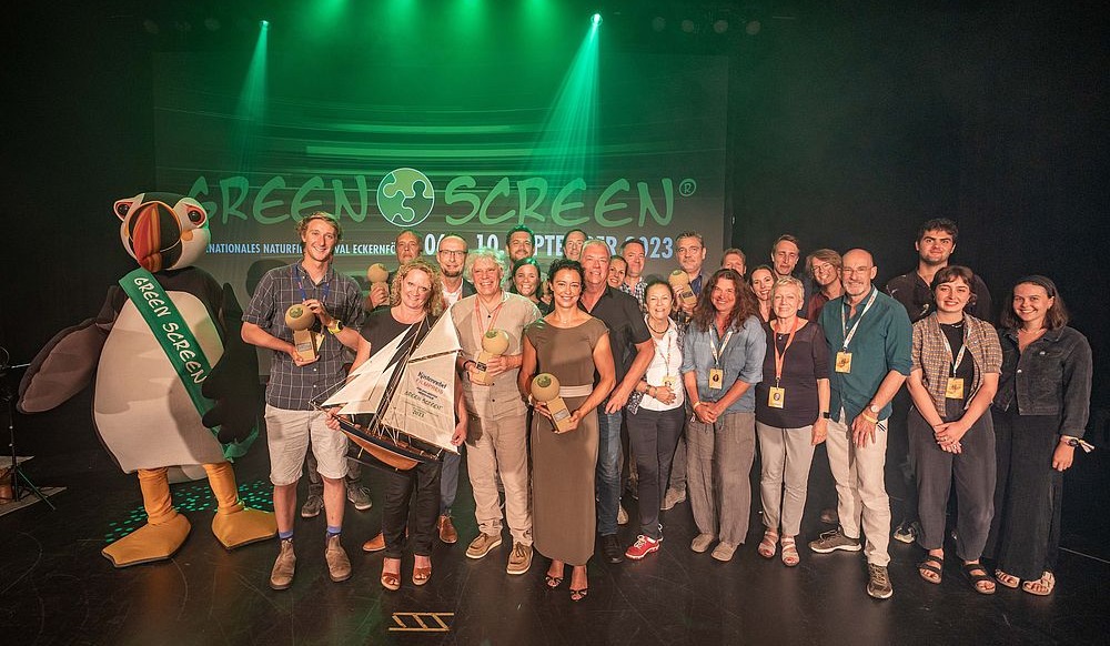 A group of people stands on stage in front of a GreenScreen 2023 banner, all smiling and holding awards. Among them, a person in a penguin costume adds a whimsical touch. The stage glows with vibrant green light.