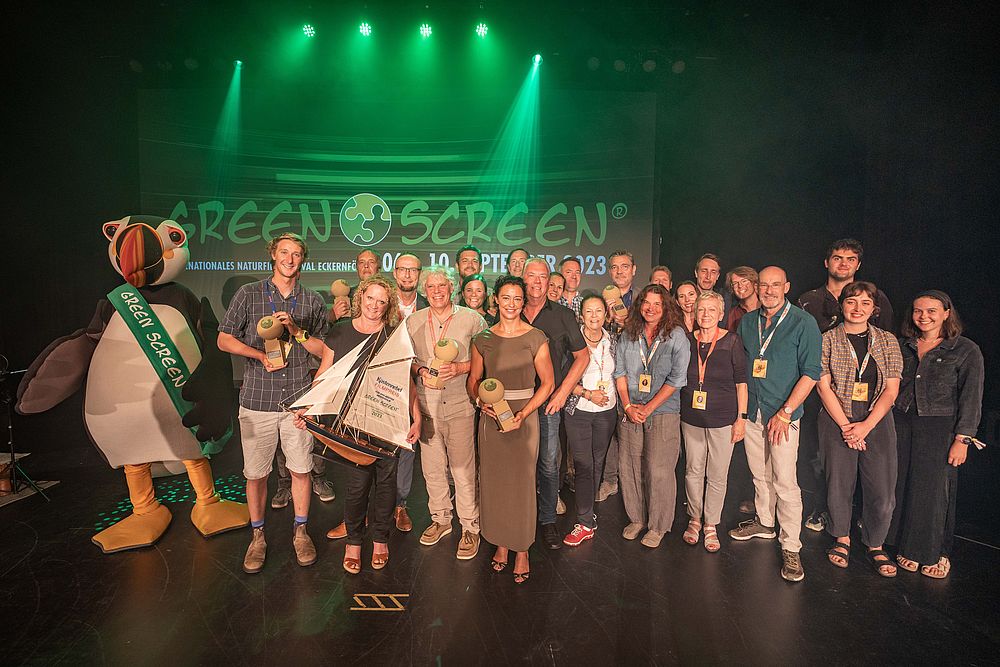 A group of people stands on stage in front of a GreenScreen 2023 banner, all smiling and holding awards. Among them, a person in a penguin costume adds a whimsical touch. The stage glows with vibrant green light.