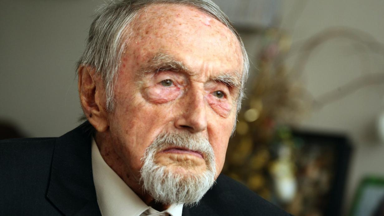 Elderly man with a white beard and mustache, reminiscent of Hans Hass, wearing a dark suit, looks thoughtfully into the distance. He has a solemn expression, with a blurred background that suggests an indoor setting.