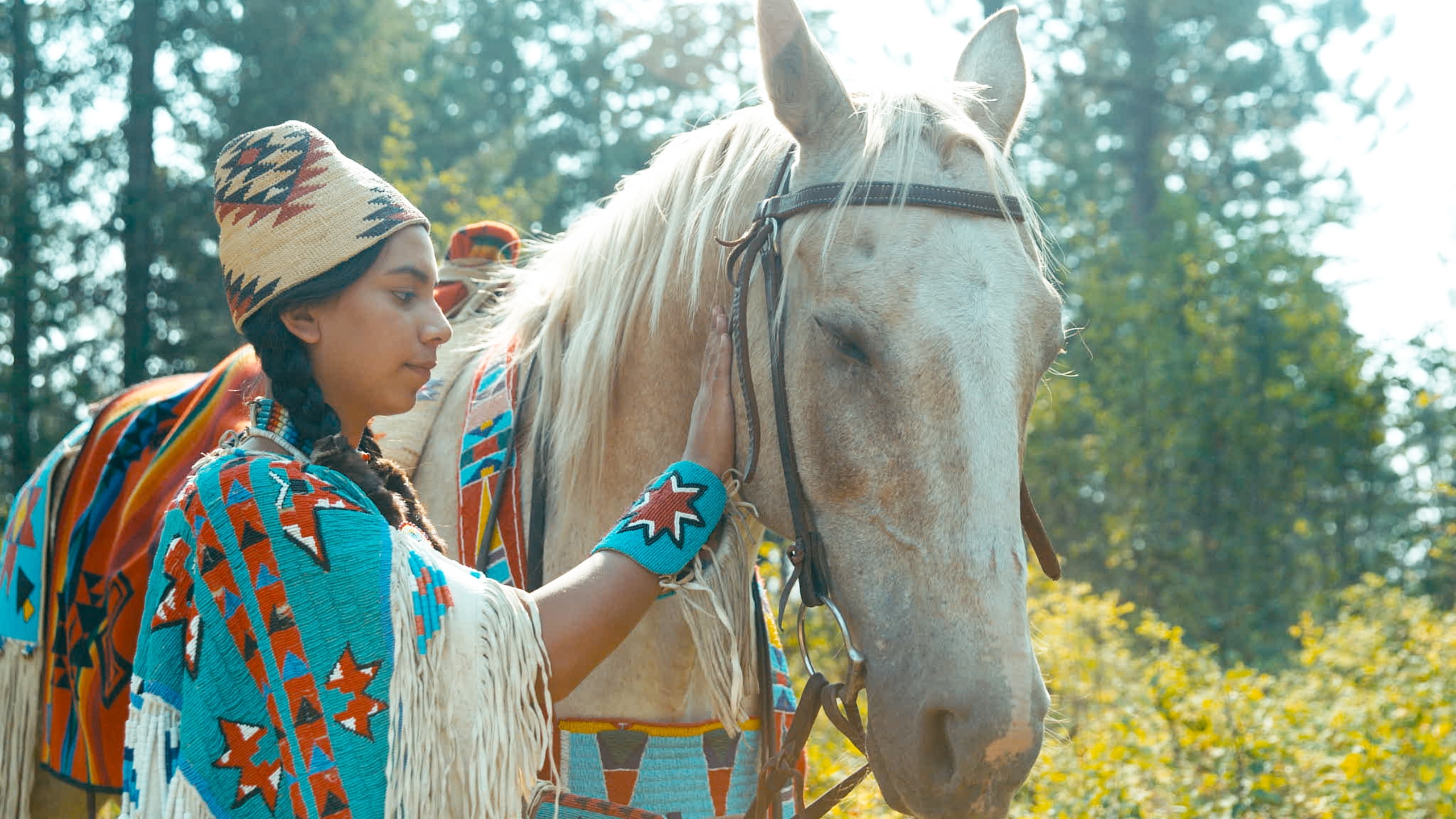 A person wearing a traditional patterned outfit and hat stands beside a white horse, symbolizing Horse Power, in a forested area. The horse is adorned with a matching colorful blanket as the sunlight filters through the trees, creating a scene reminiscent of American Legacies.