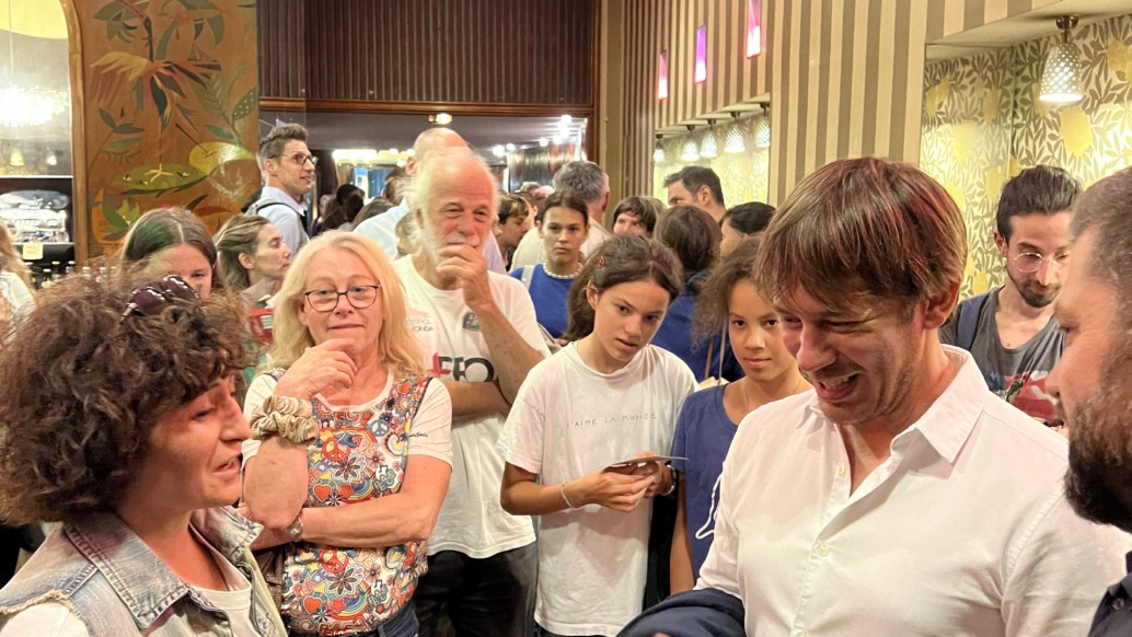 A crowded room buzzes with chatter and smiles as a man in a white shirt engages with others. The setting, perfect for feature premieres, boasts ornate wallpaper and warm lighting. A mix of men, women, and children are present, some holding phones or wearing glasses.