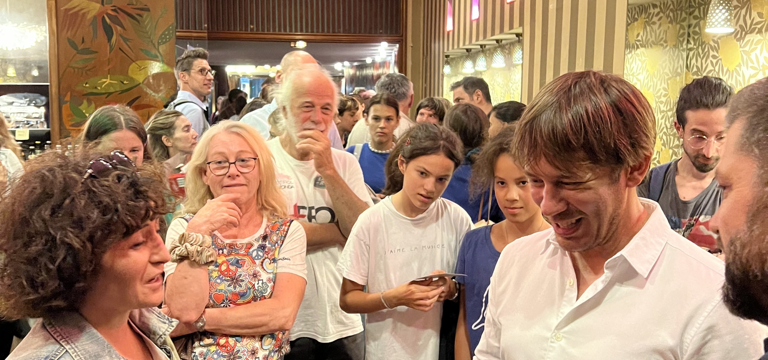 A crowded room buzzes with chatter and smiles as a man in a white shirt engages with others. The setting, perfect for feature premieres, boasts ornate wallpaper and warm lighting. A mix of men, women, and children are present, some holding phones or wearing glasses.