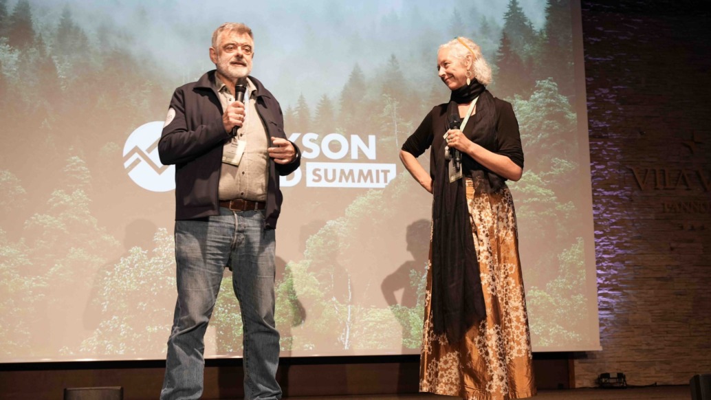 An older man and woman stand on stage holding microphones at the Jackson Wild Summit 2022. The backdrop features a logo and image of a lush forest. The man wears a jacket and jeans while the woman sports a long skirt and scarf, embodying TMS Productions' commitment to environmental awareness.