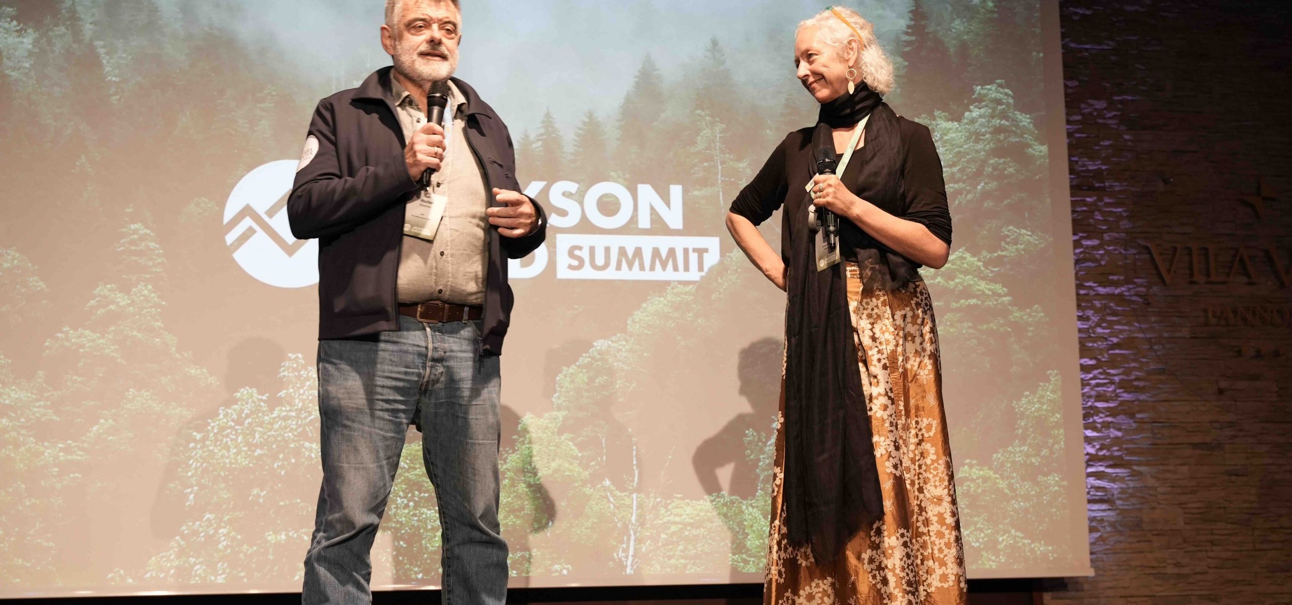 An older man and woman stand on stage holding microphones at the Jackson Wild Summit 2022. The backdrop features a logo and image of a lush forest. The man wears a jacket and jeans while the woman sports a long skirt and scarf, embodying TMS Productions' commitment to environmental awareness.