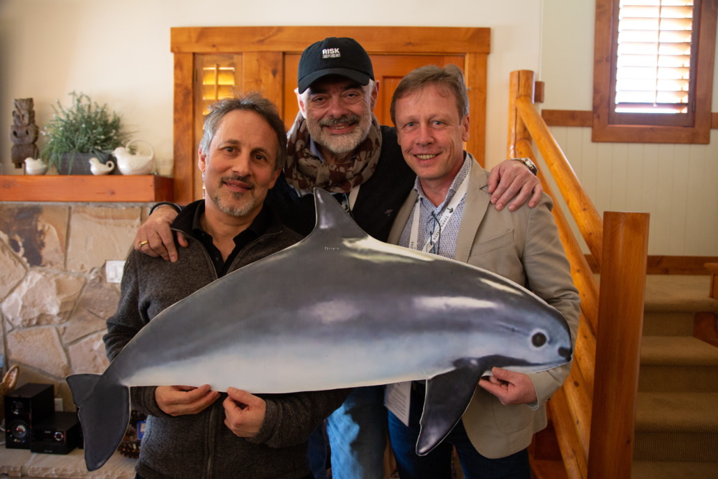 Three men stand in a room with wooden decor, smiling and holding a large cutout of a dolphin. The man in the middle, wearing a hat and scarf, exudes casual charm. This friendly gathering evokes memories of the Sundance Premiere of "Sea of Shadows.