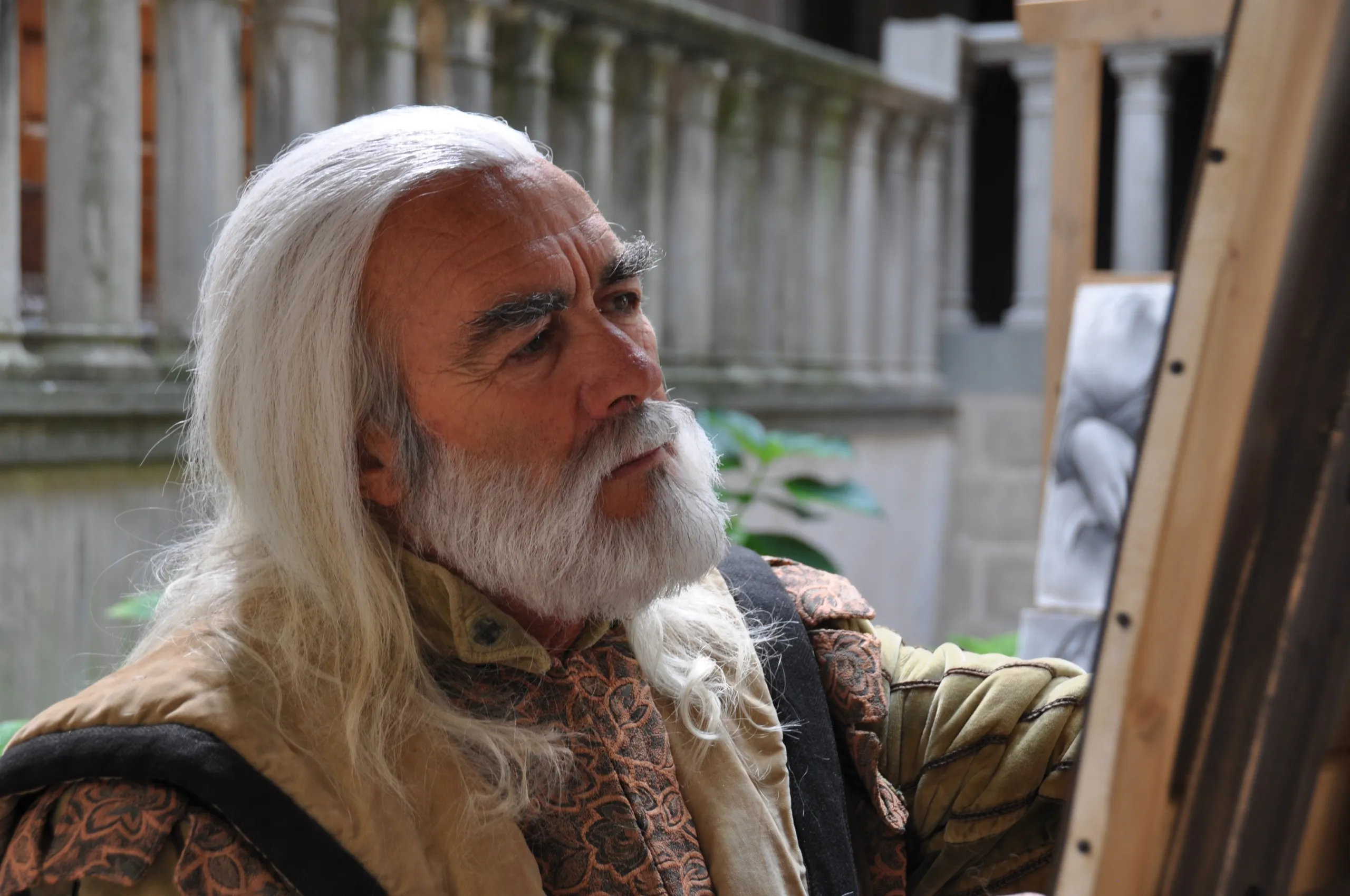 An elderly man with long white hair and a beard is painting at an easel outdoors, perhaps capturing his own version of the Mona Lisa. He is wearing a patterned shirt and vest, with a stone railing and part of a building visible in the background.