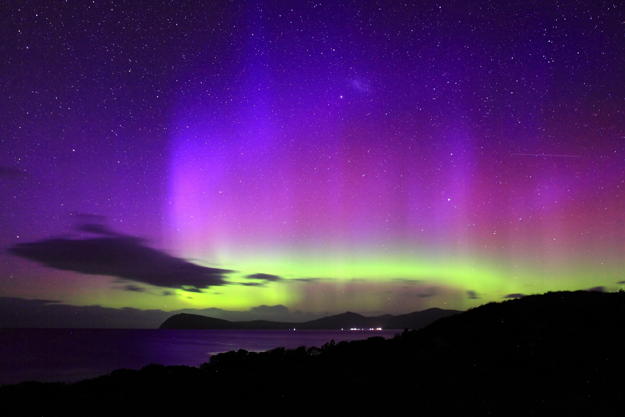 A stunning display of the aurora borealis casts a spell of nature, lighting up the night sky with gradients of purple, pink, and green. Below, a serene coastline and dark silhouetted hills lie under the starlit sky, capturing magic moments in time.