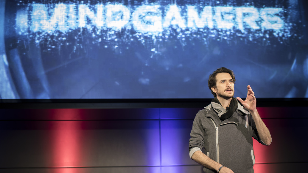 A person stands on stage delivering a captivating presentation with a MindGamers backdrop. The setting, illuminated in red and blue lights, creates an electrifying atmosphere blending science and entertainment. The speaker gestures smoothly with one hand, dressed in casual attire.