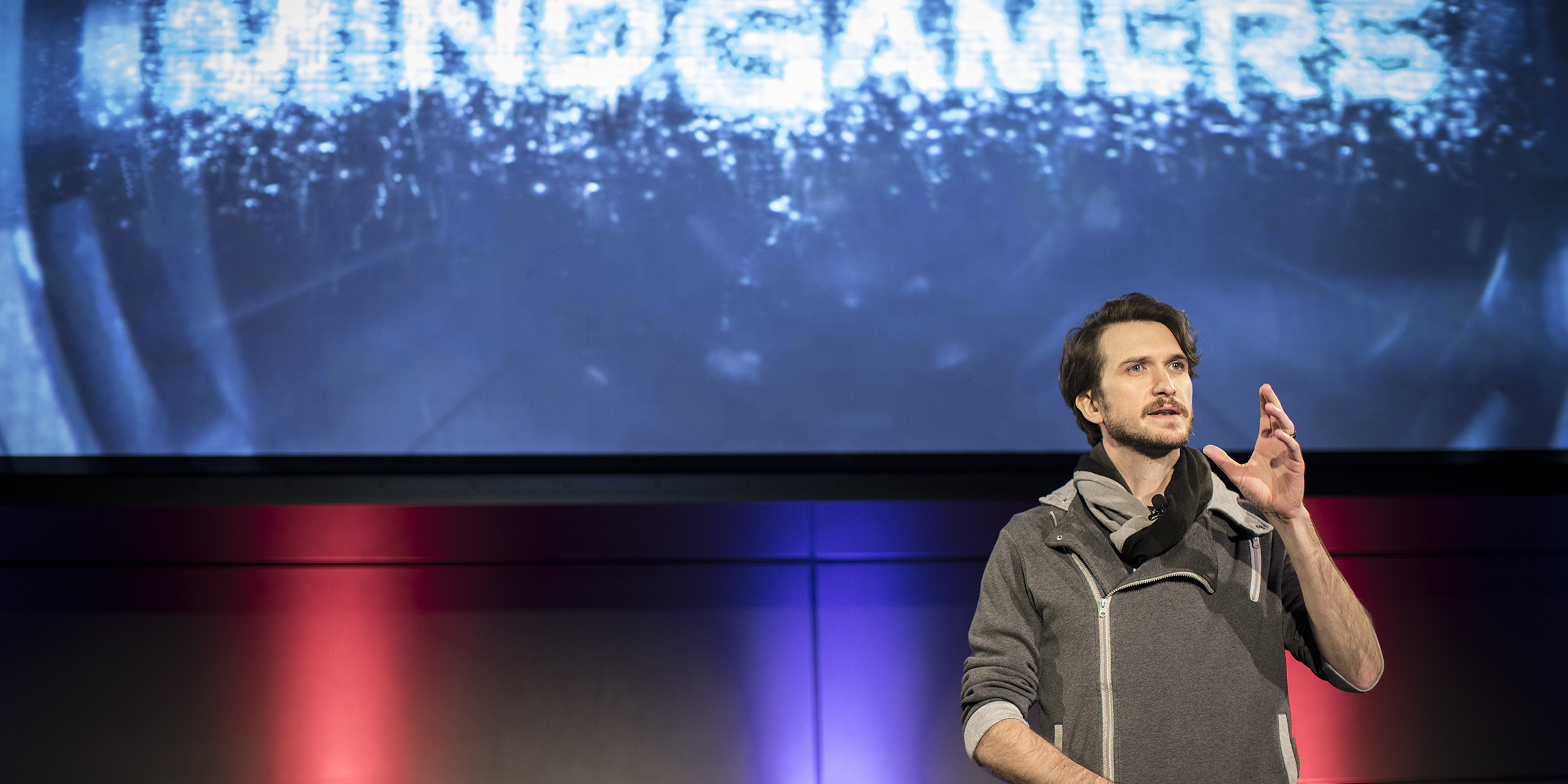 A person stands on stage delivering a captivating presentation with a MindGamers backdrop. The setting, illuminated in red and blue lights, creates an electrifying atmosphere blending science and entertainment. The speaker gestures smoothly with one hand, dressed in casual attire.