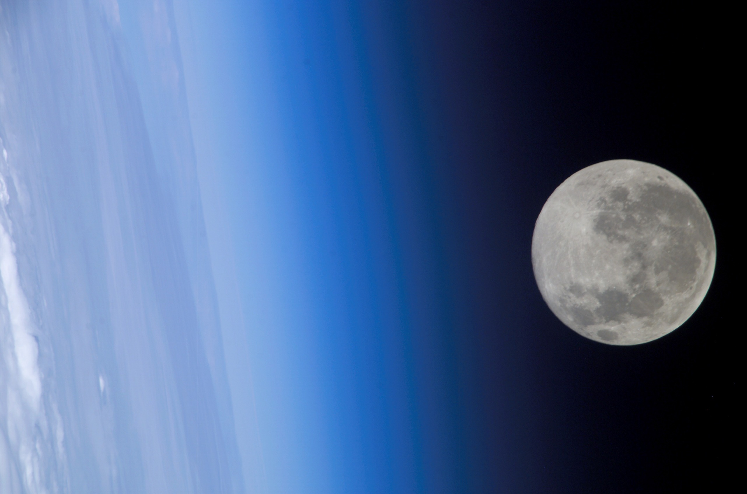 A full moon is visible against the dark sky, with Earth's atmosphere casting a gradient of blue shades on the left. The moon's glow enhances the contrasting colors, underscoring the celestial scene.