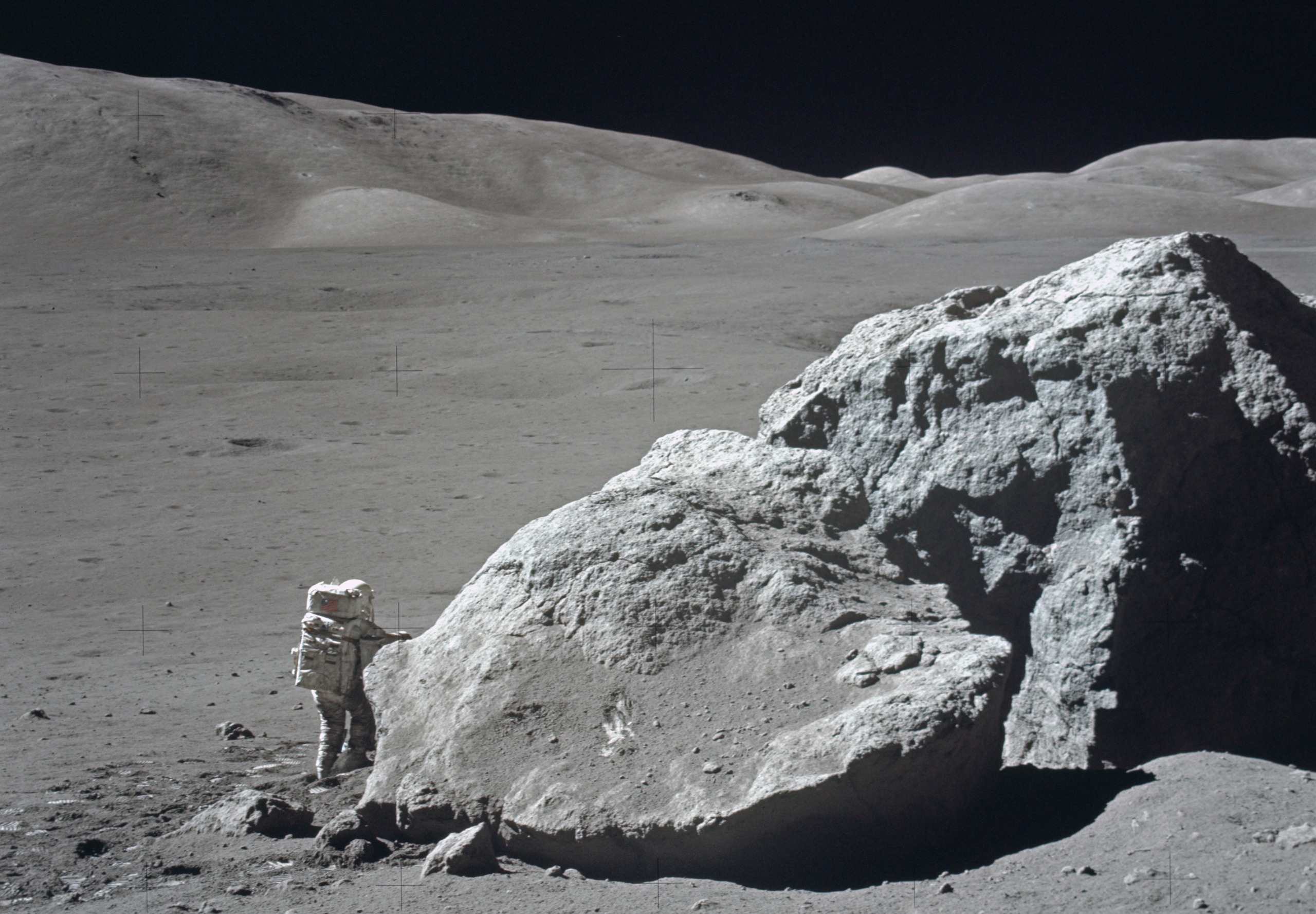 An astronaut in a spacesuit stands beside a large boulder on the moon's surface under a dark sky. The lunar landscape is barren, with gentle hills in the background.