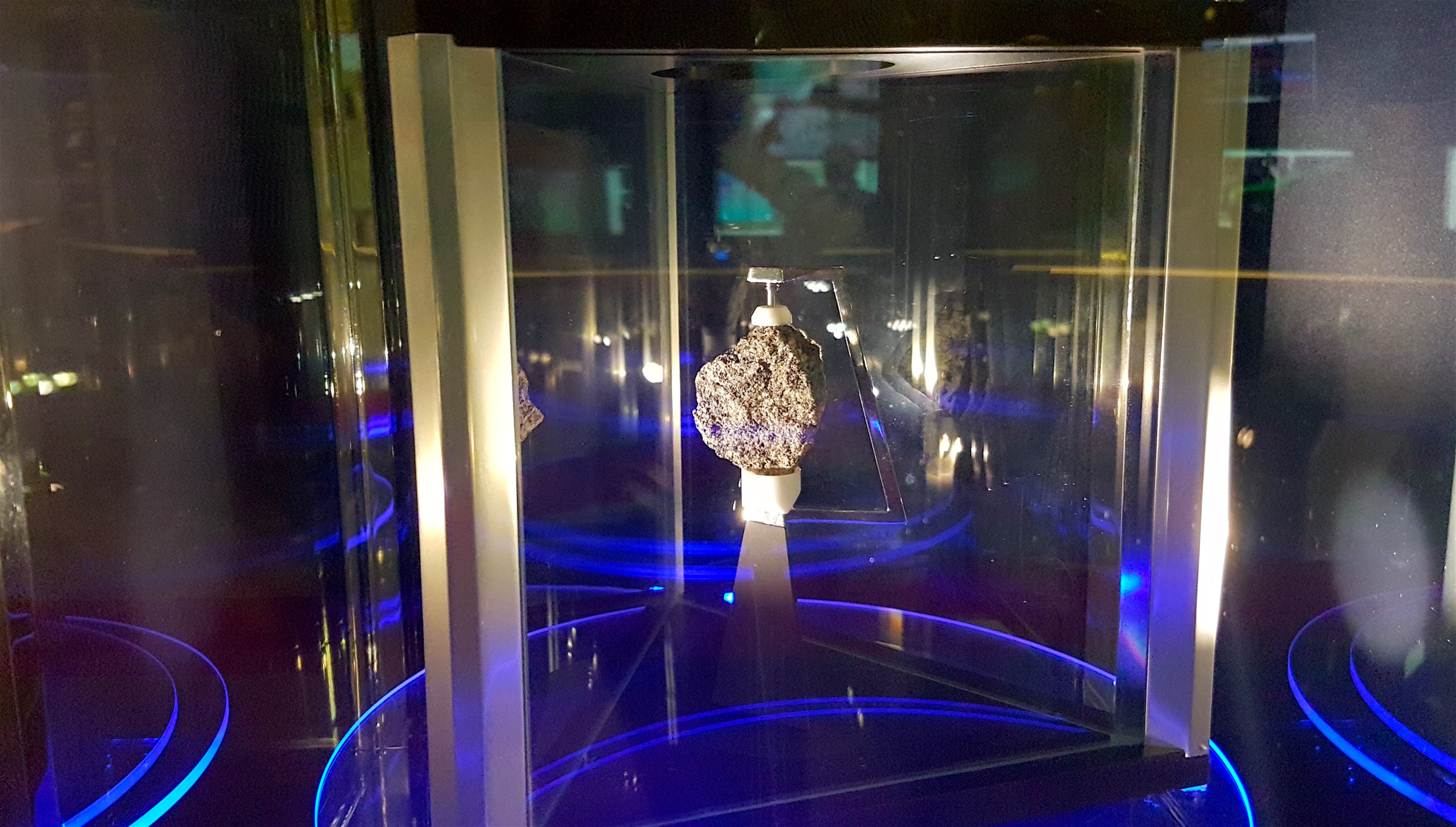 A meteorite, akin to a relic from the moon, glows in a glass case illuminated by blue lights. Its rough, textured surface is mounted on a white stand, commanding attention at the exhibit's center.