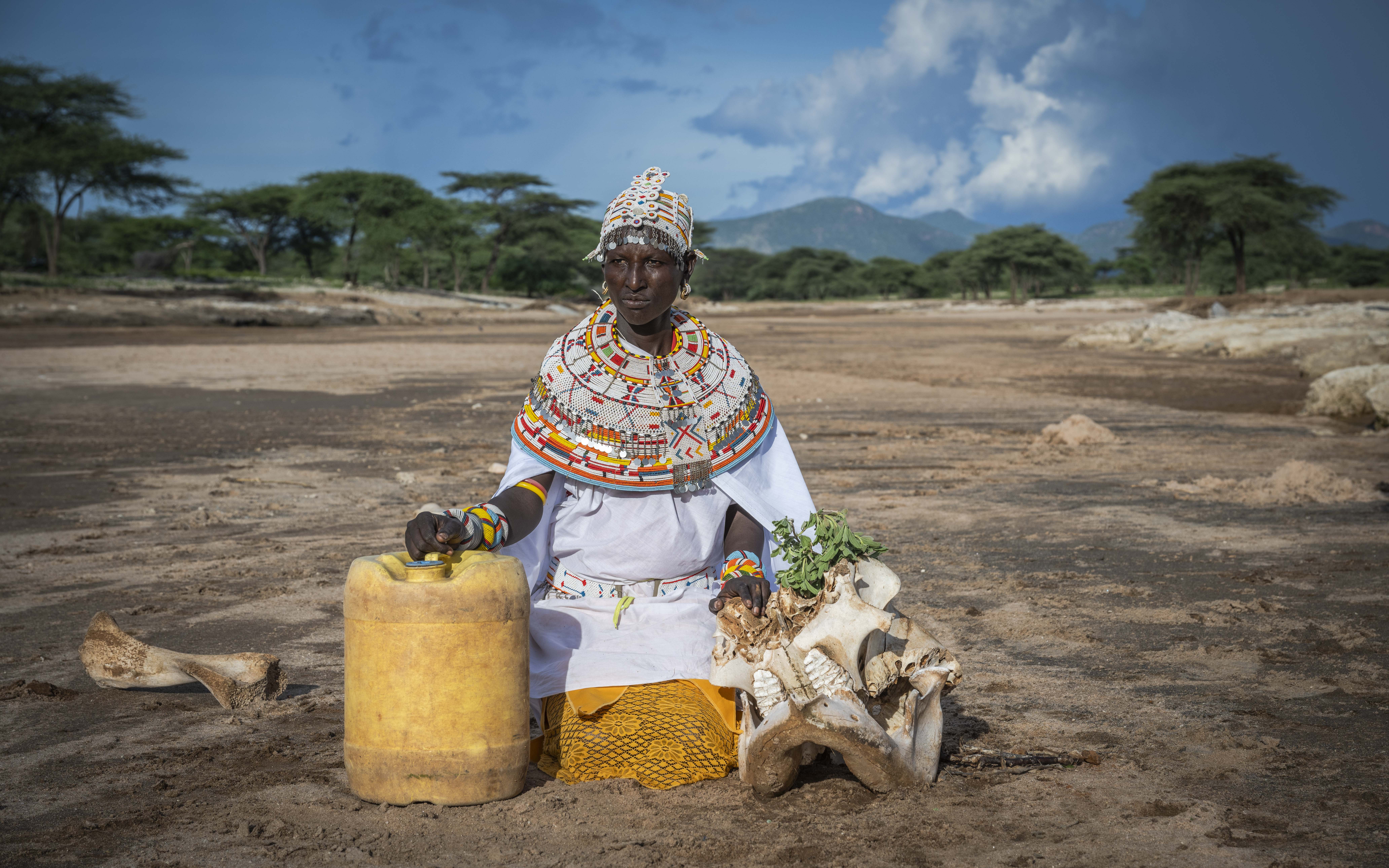 A person in traditional attire sits on dry, cracked earth with a yellow jerrycan and animal skulls, evoking scenes from Ovid's *Metamorphoses*. The landscape mirrors ancient tales, with bushes and mountains in the background under a cloudy blue sky reminiscent of Roman mythology.