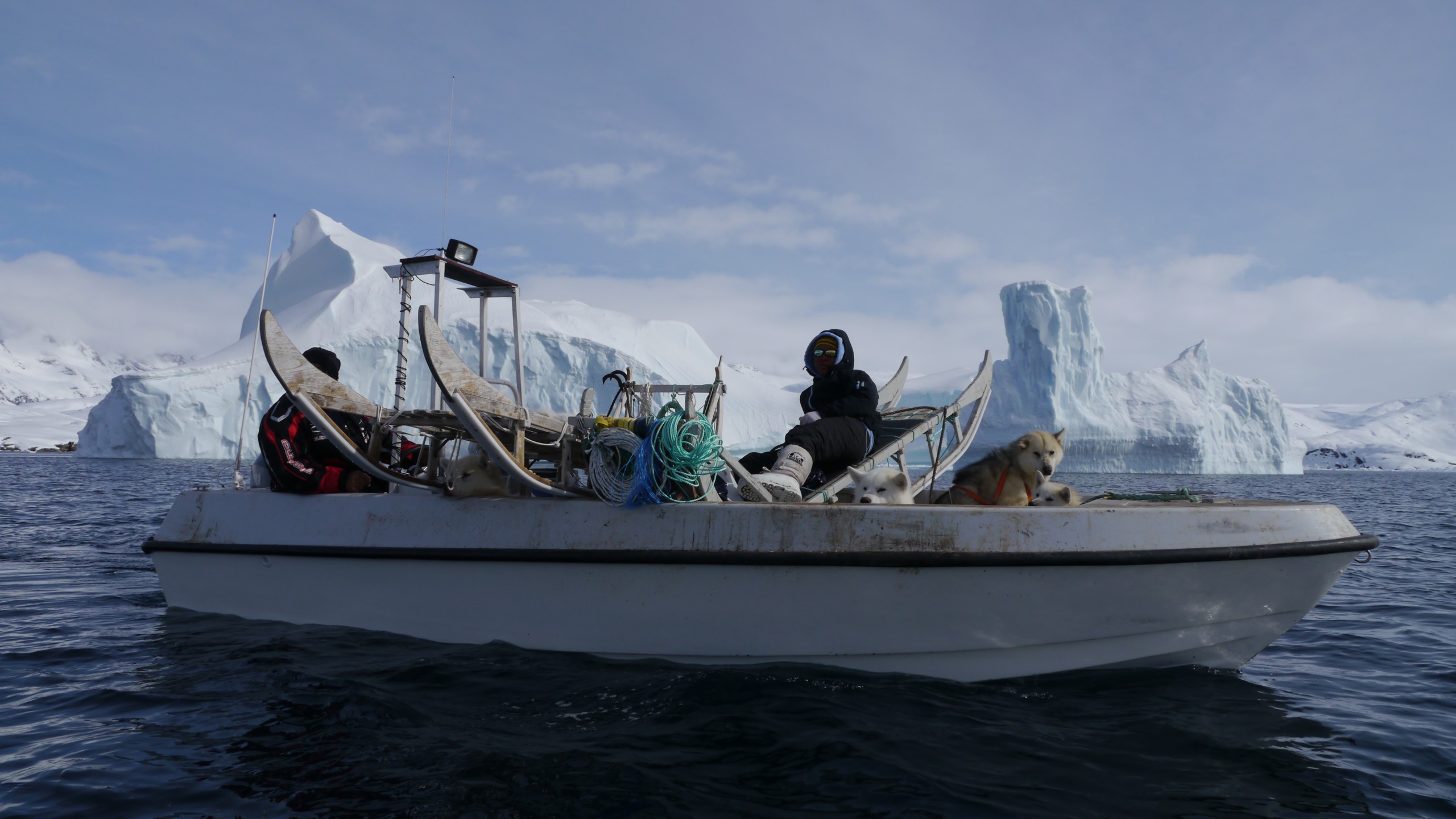 A small boat with a person and a dog on board floats in icy waters, embodying the essence of being next to nature. The boat, laden with supplies, is surrounded by large icebergs under a clear blue sky, while snow-covered mountains loom majestically in the background.