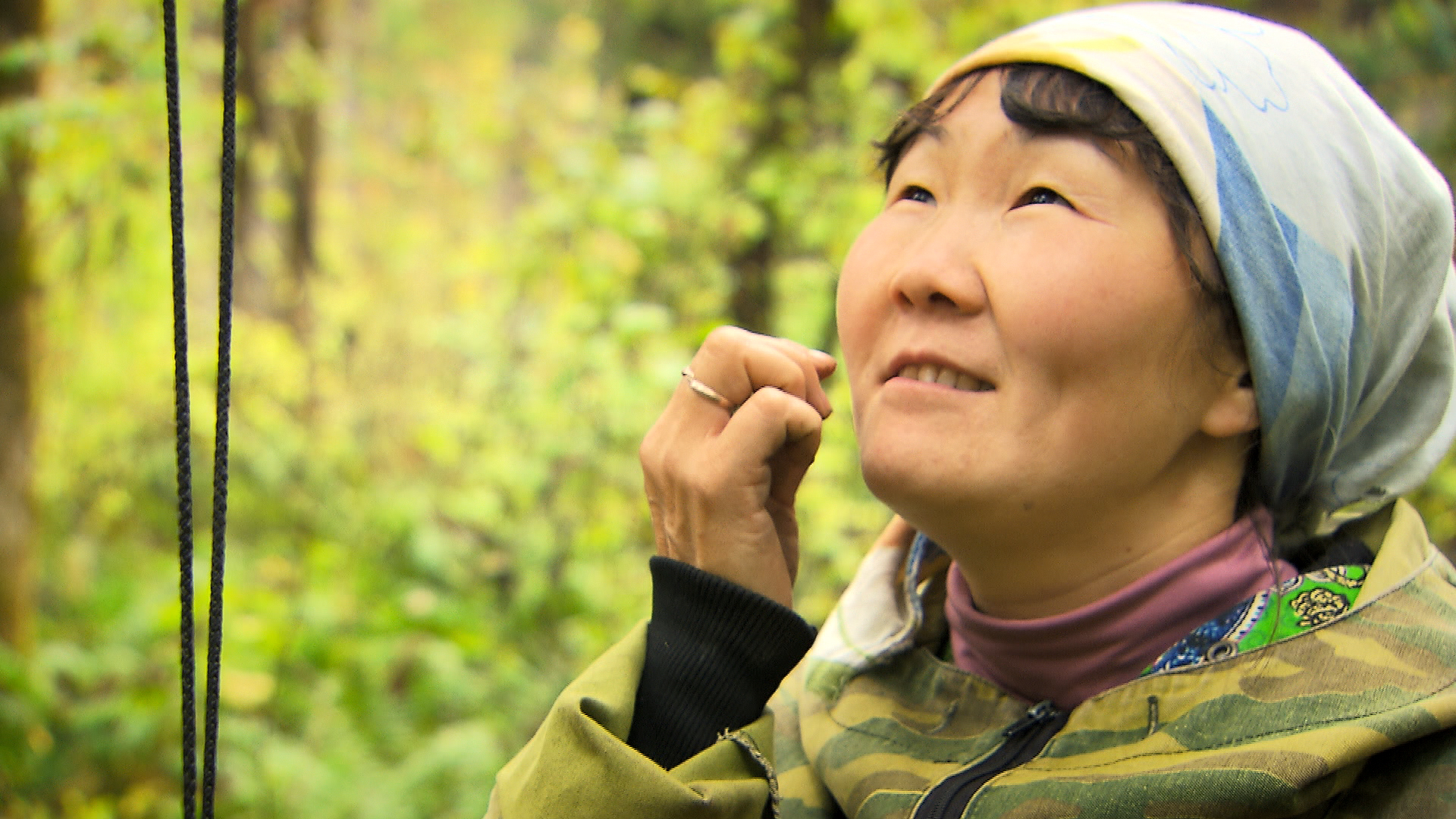 A person wearing a headscarf and a camouflage jacket looks upward with a serene expression in a lush, green forest setting. They appear to be gently touching their cheek, perfectly attuned with the tranquility of nature.