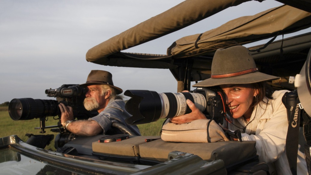 Two people wearing hats and using large cameras with telephoto lenses sit in an open safari vehicle, as if capturing footage worthy of the Nature Film Awards. They appear focused on photographing wildlife in a grassy landscape under a clear sky.