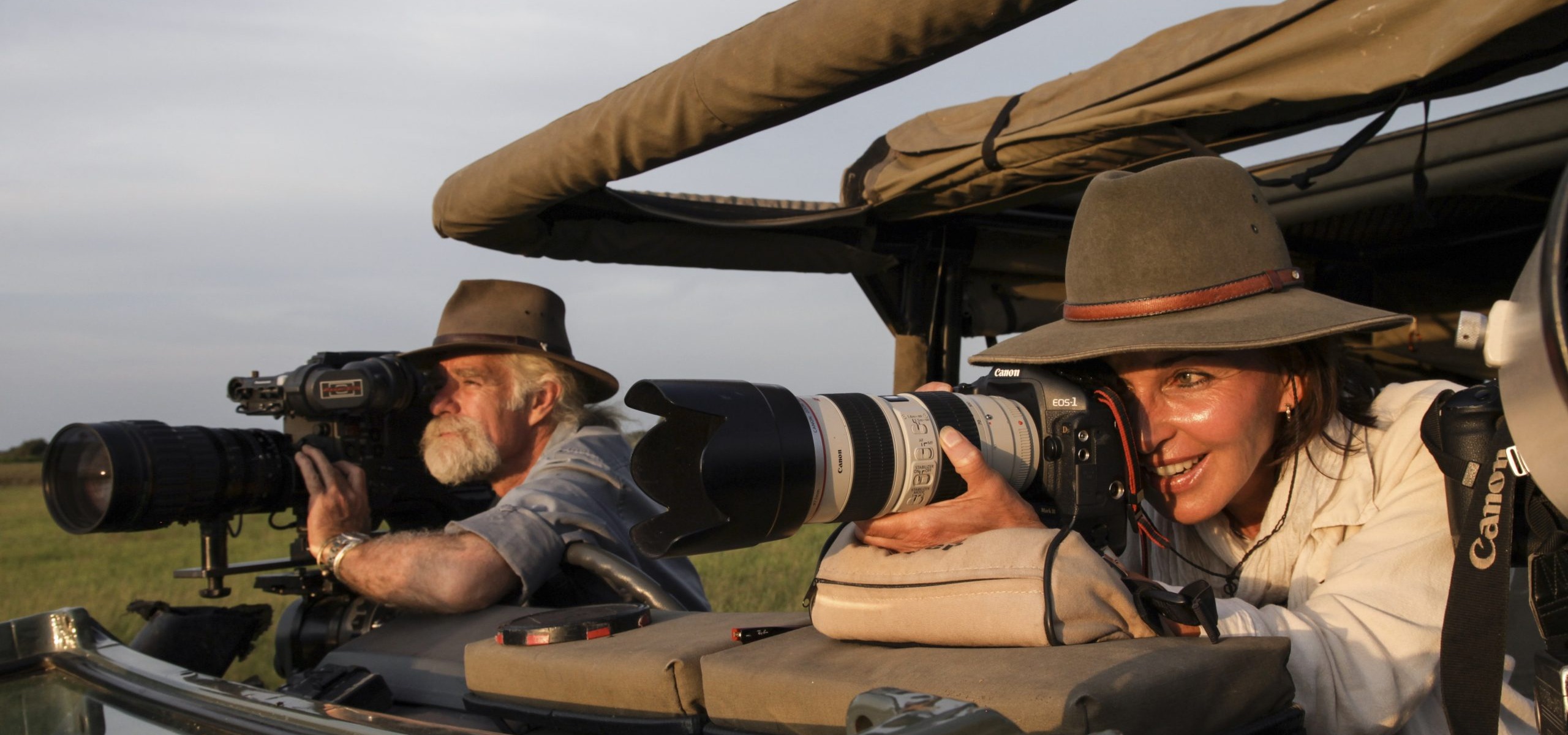 Two people wearing hats and using large cameras with telephoto lenses sit in an open safari vehicle, as if capturing footage worthy of the Nature Film Awards. They appear focused on photographing wildlife in a grassy landscape under a clear sky.