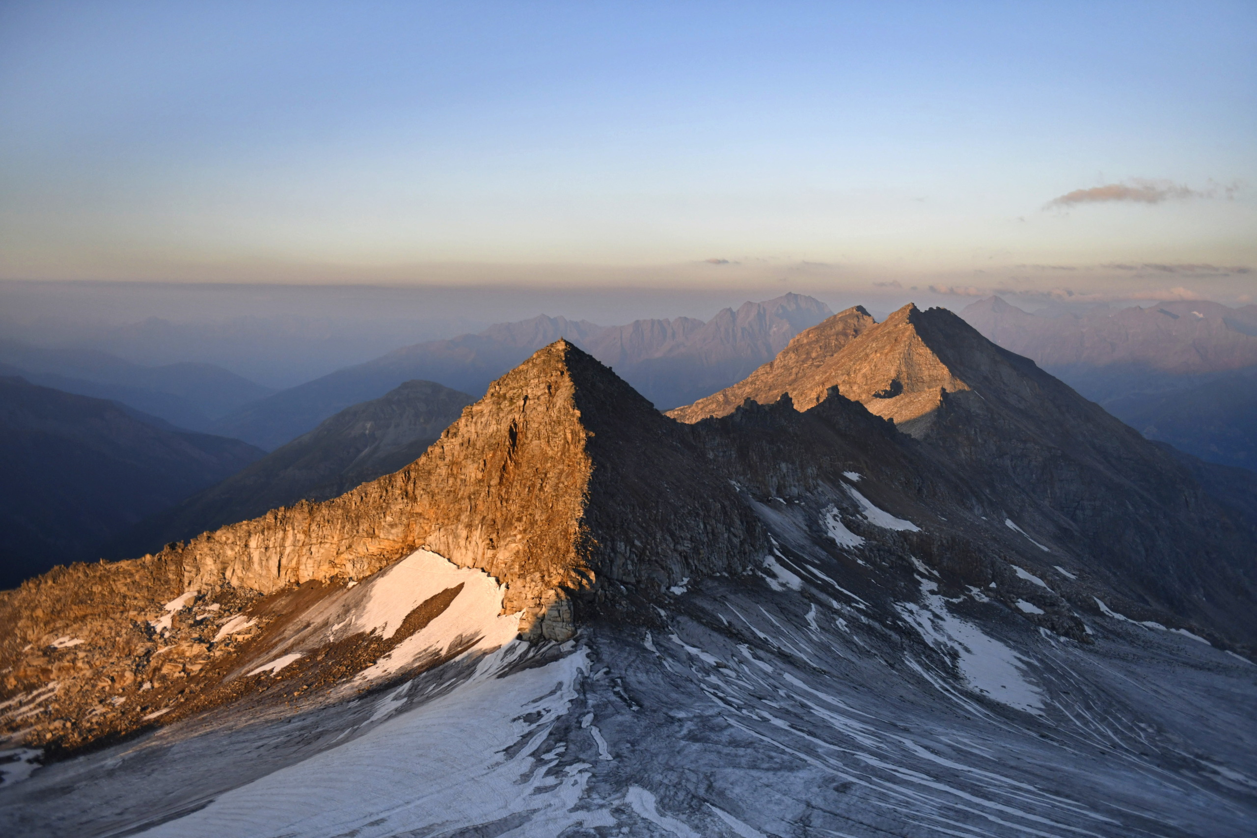 A mountainous landscape illuminated by sunset casts warm light on the rugged peaks of the Alps, with patches of snow. The foreground features rocky terrain, while distant mountains fade into a hazy blue horizon under a serene sky.