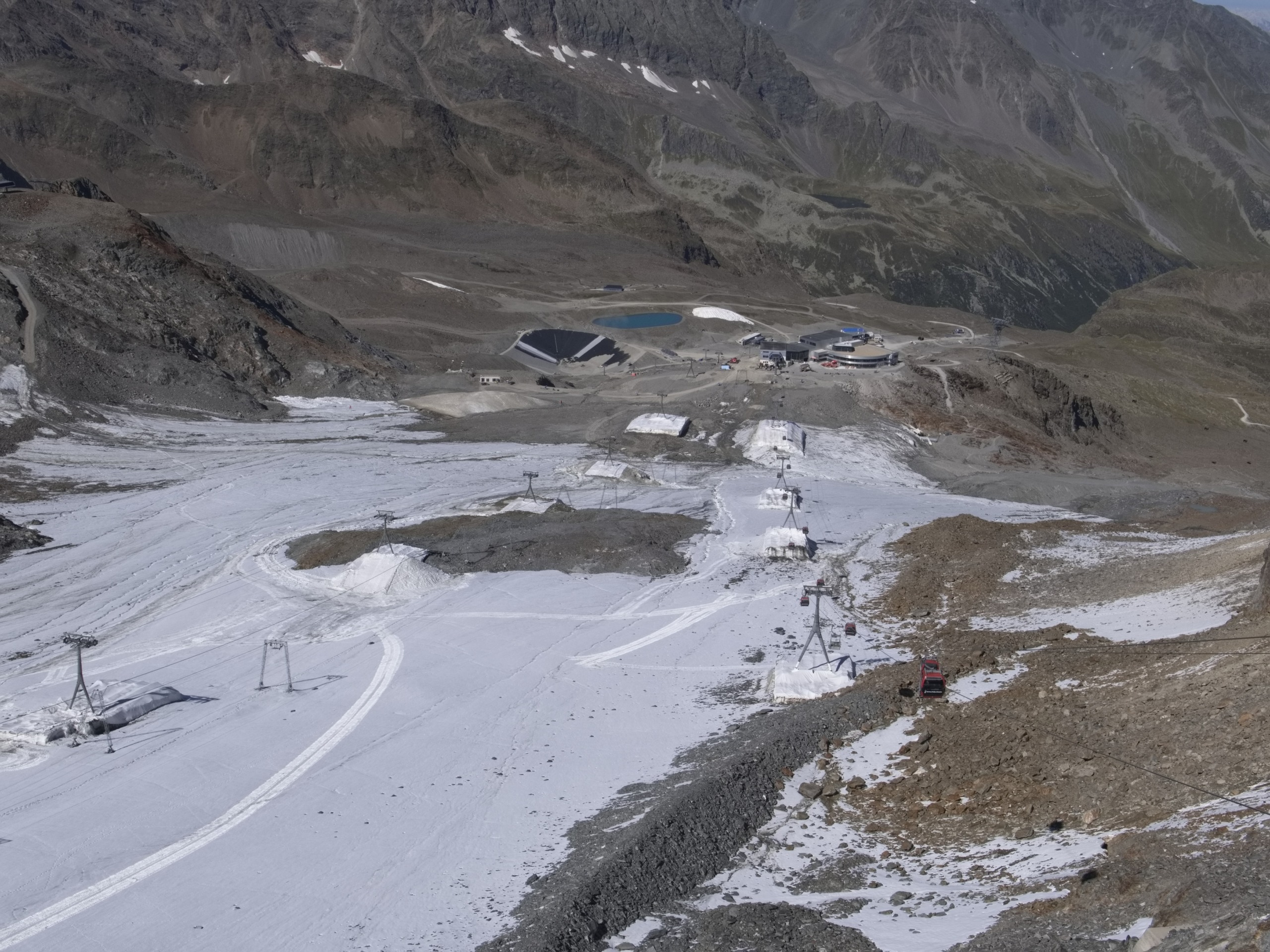 A mountain landscape featuring a ski area with ski lifts on a partially snow-covered slope in the majestic Alps. The background reveals rugged mountains and a small building complex. The sky is clear, with patches of green vegetation dotting the hillsides.