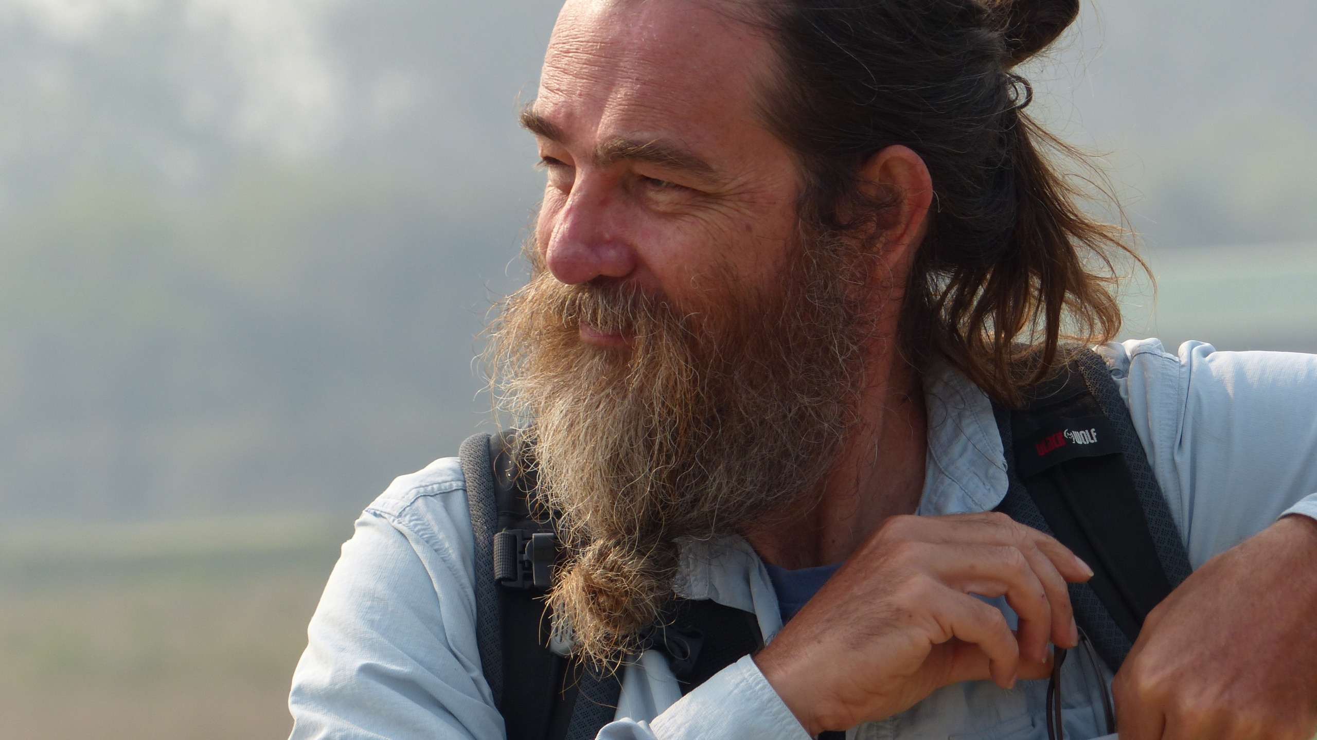 A person with long hair and a beard stands outdoors, wearing a light-colored shirt and a backpack. They appear thoughtful, perhaps contemplating secrets, as they look to the side with a slight smile. The background is blurred, suggesting a natural setting.