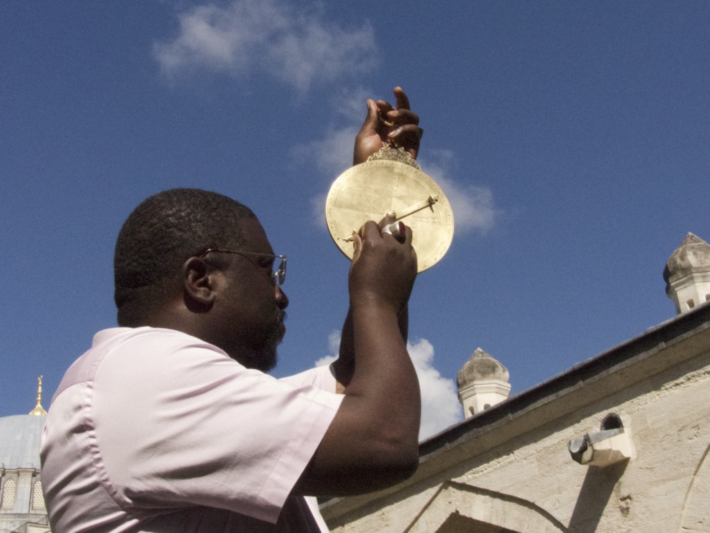 A Star Chaser holds and examines an astrolabe against a clear blue sky, with a building featuring domes and minarets in the background, reminiscent of Senegal's vibrant architectural heritage.