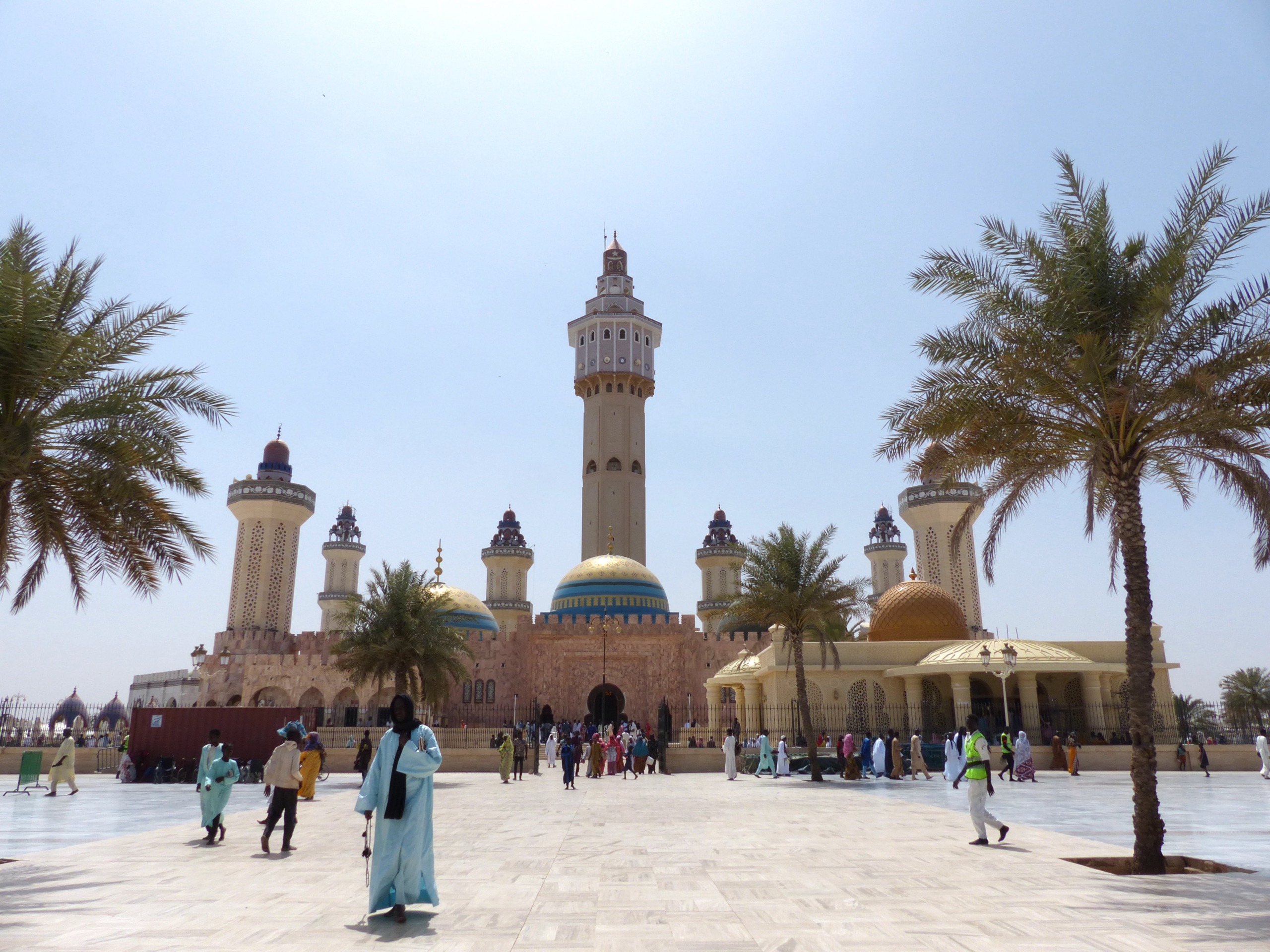 Under the clear blue sky, the scenic view of a large mosque with multiple minarets and domes is mesmerizing. In Senegal, people in colorful traditional attire stroll across the spacious courtyard lined with palm trees, resembling Star Chasers seeking serenity.