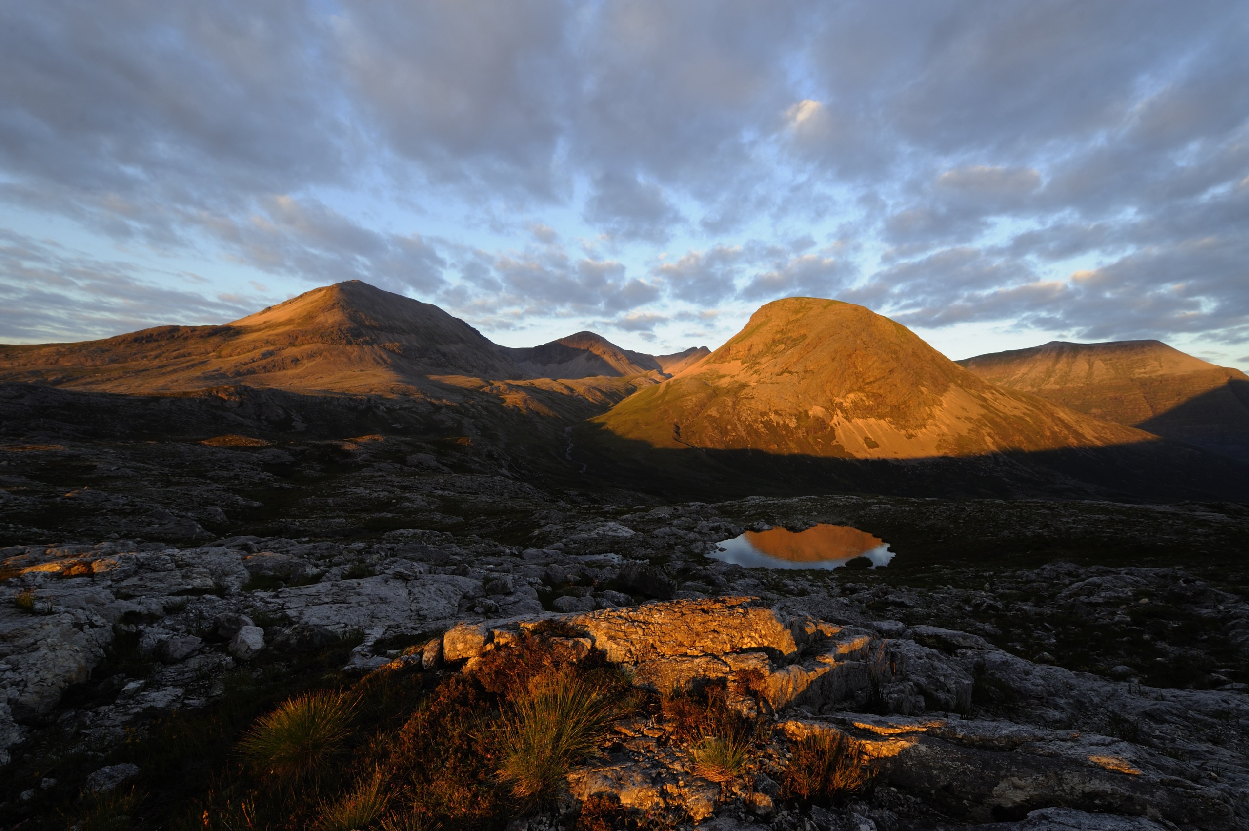 A landscape in Scotland features rugged mountains under a partly cloudy sky at sunset. The warm light casts shadows and highlights the rocky terrain. A small reflective pond is in the foreground, mirroring the golden hues of this breathtaking scene.