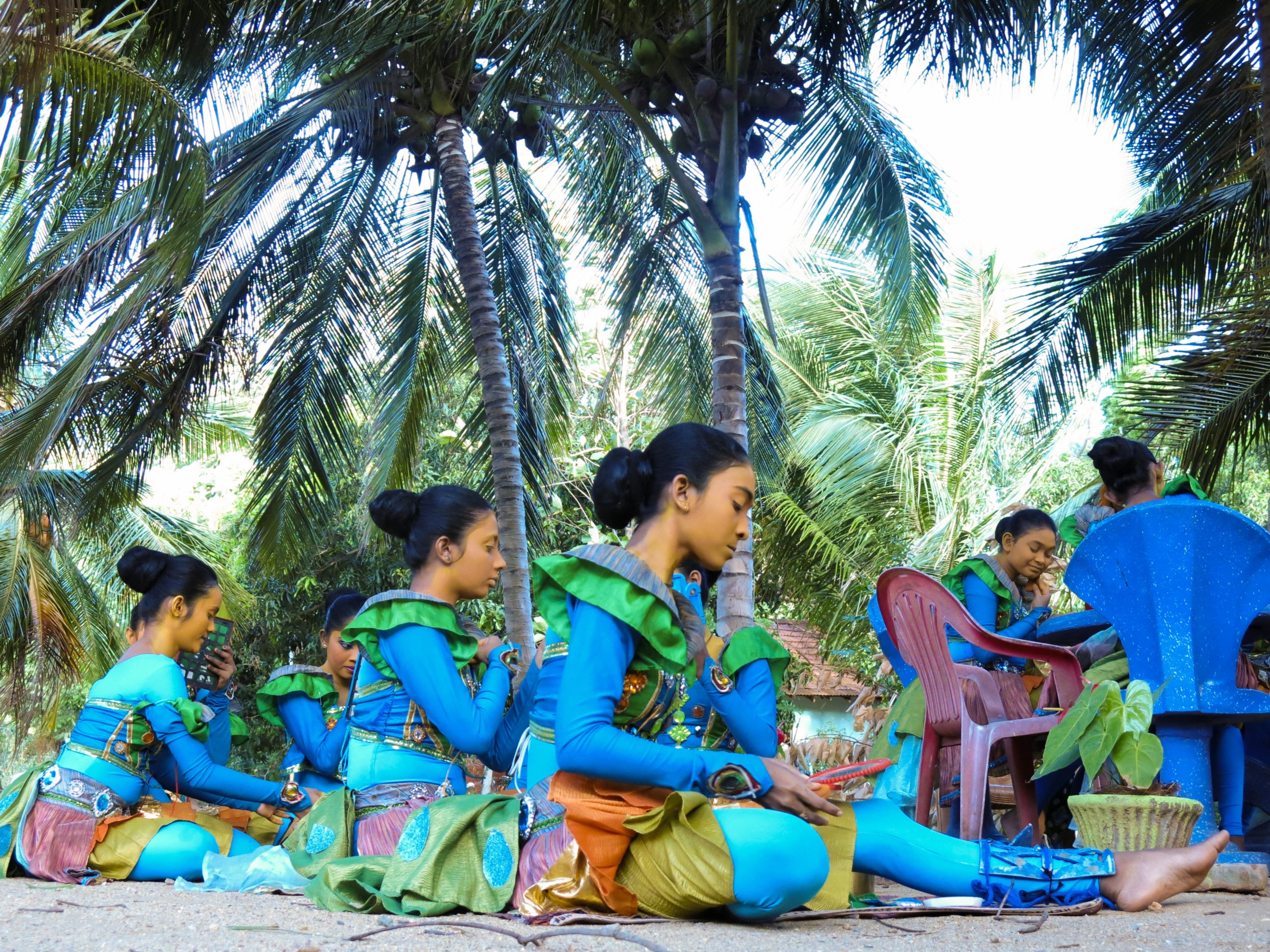 A group of young people in vibrant blue and green costumes sits under tall palm trees in Sri Lanka. Some are seated on red plastic chairs, while others relax on the sandy ground, one holding a phone.