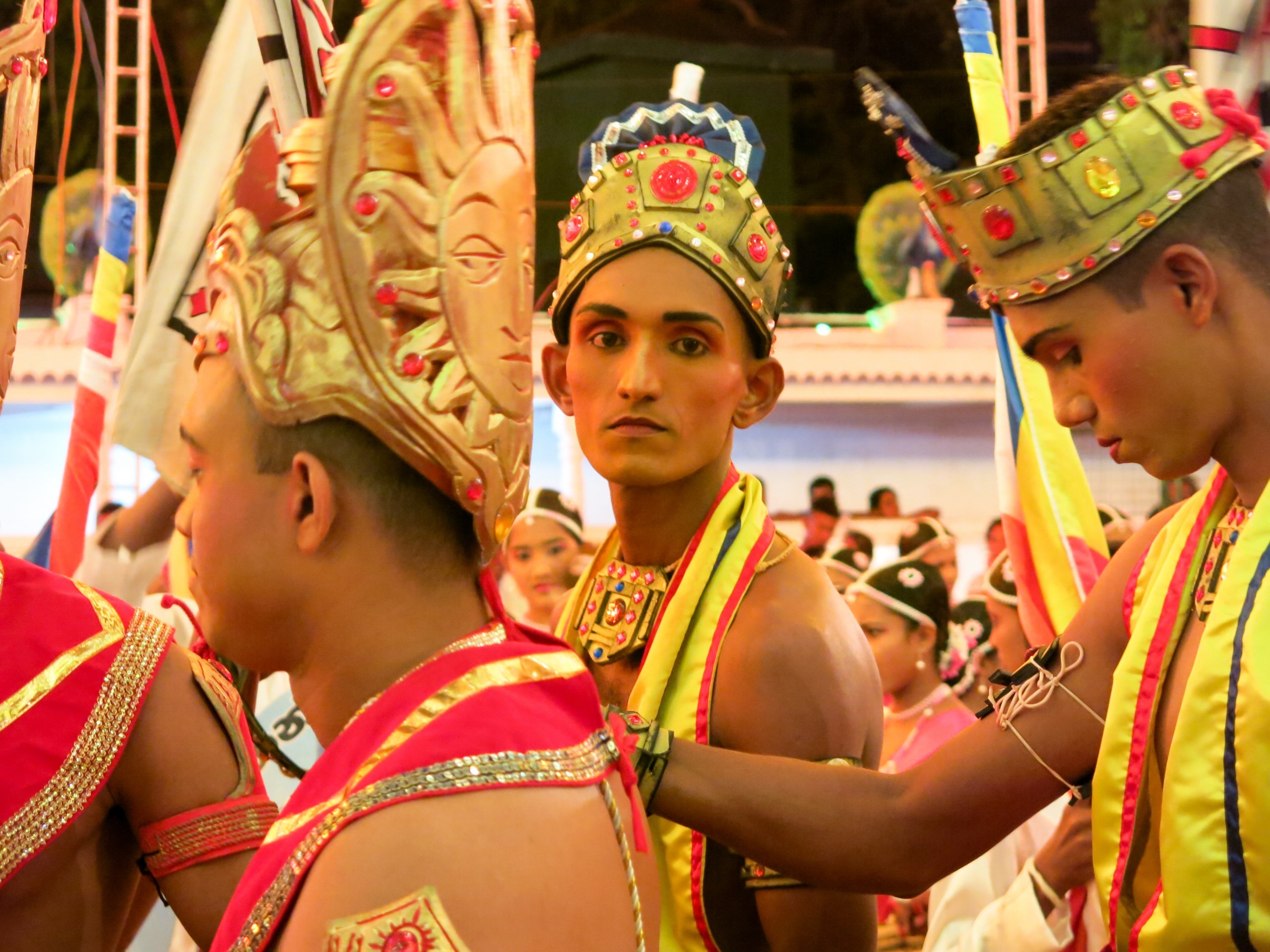 A group of people in traditional, colorful costumes and ornate headdresses participate in a cultural event reminiscent of Sri Lanka's rich heritage. They stand closely together, with vibrant decorations enhancing the festive atmosphere.