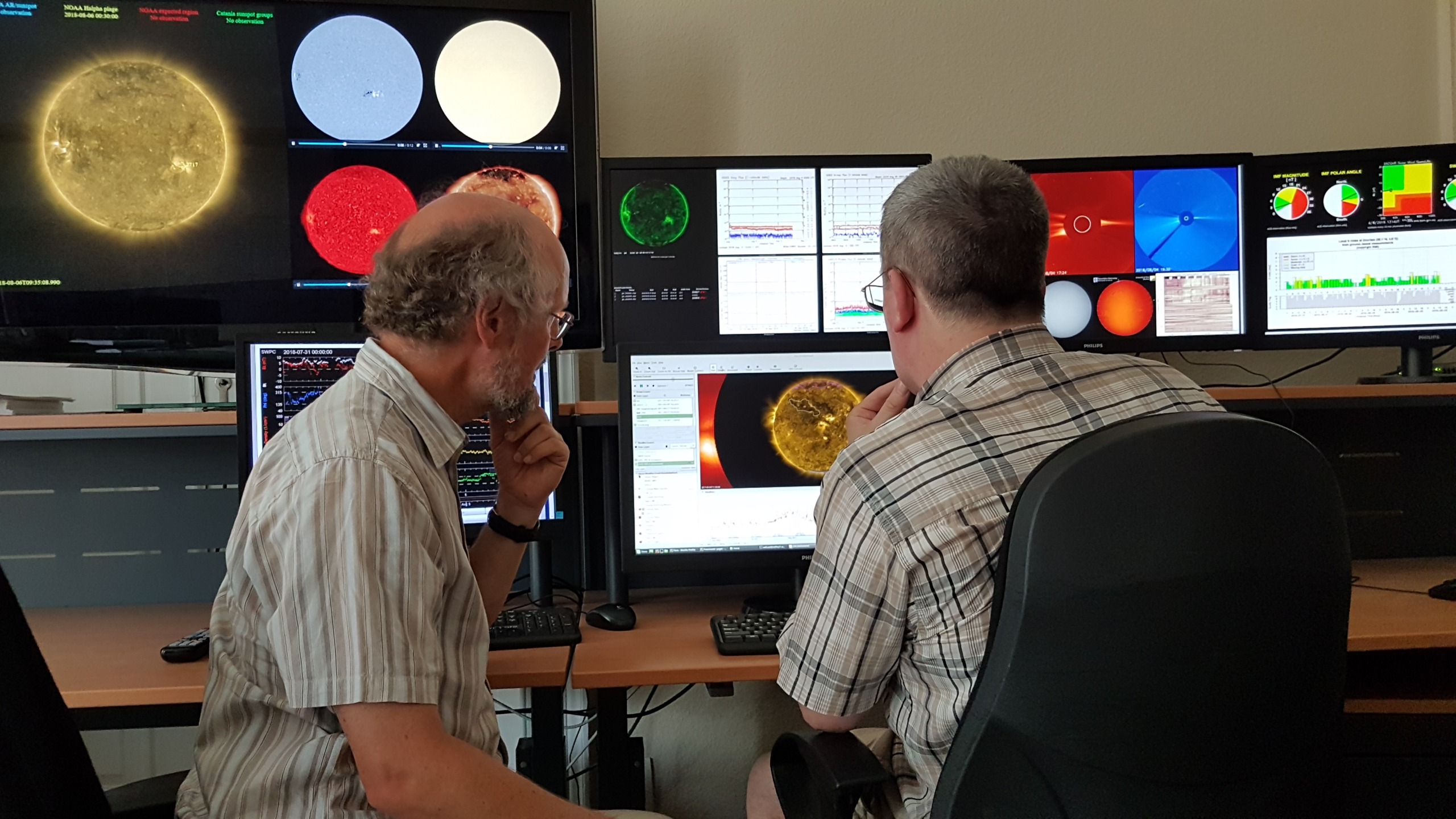 In a control room beneath the vast sky, two men intently analyze data on multiple screens, each displaying vivid charts and images of the sun. The inferno-like hues command their focused attention as they decipher the celestial information.