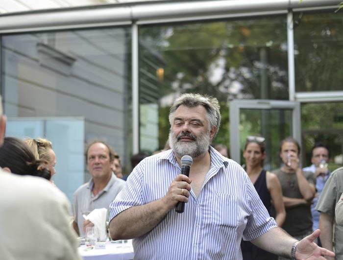 A bearded man holds a microphone, speaking to a group of people at the 2013 Summer Party. The crowd listens attentively, some standing and others seated, as they enjoy the TMFS event. Glass doors and greenery provide a serene backdrop to this lively gathering.