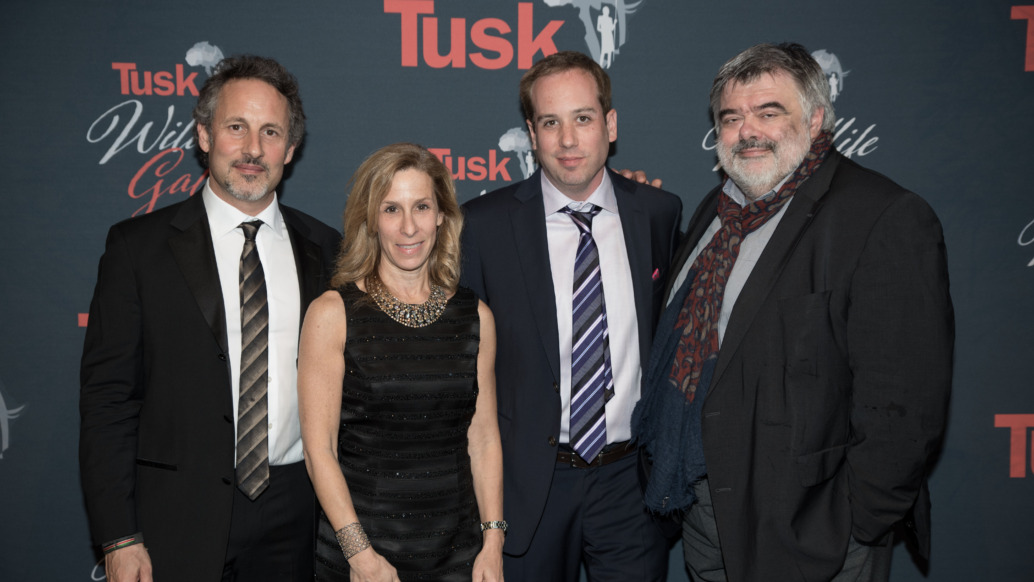 Four people pose together in formal attire against a backdrop featuring the word Tusk and an outline of Africa. Three men in suits and a woman in a black dress smile at the camera, celebrating their honor with the Game-Changer Award.