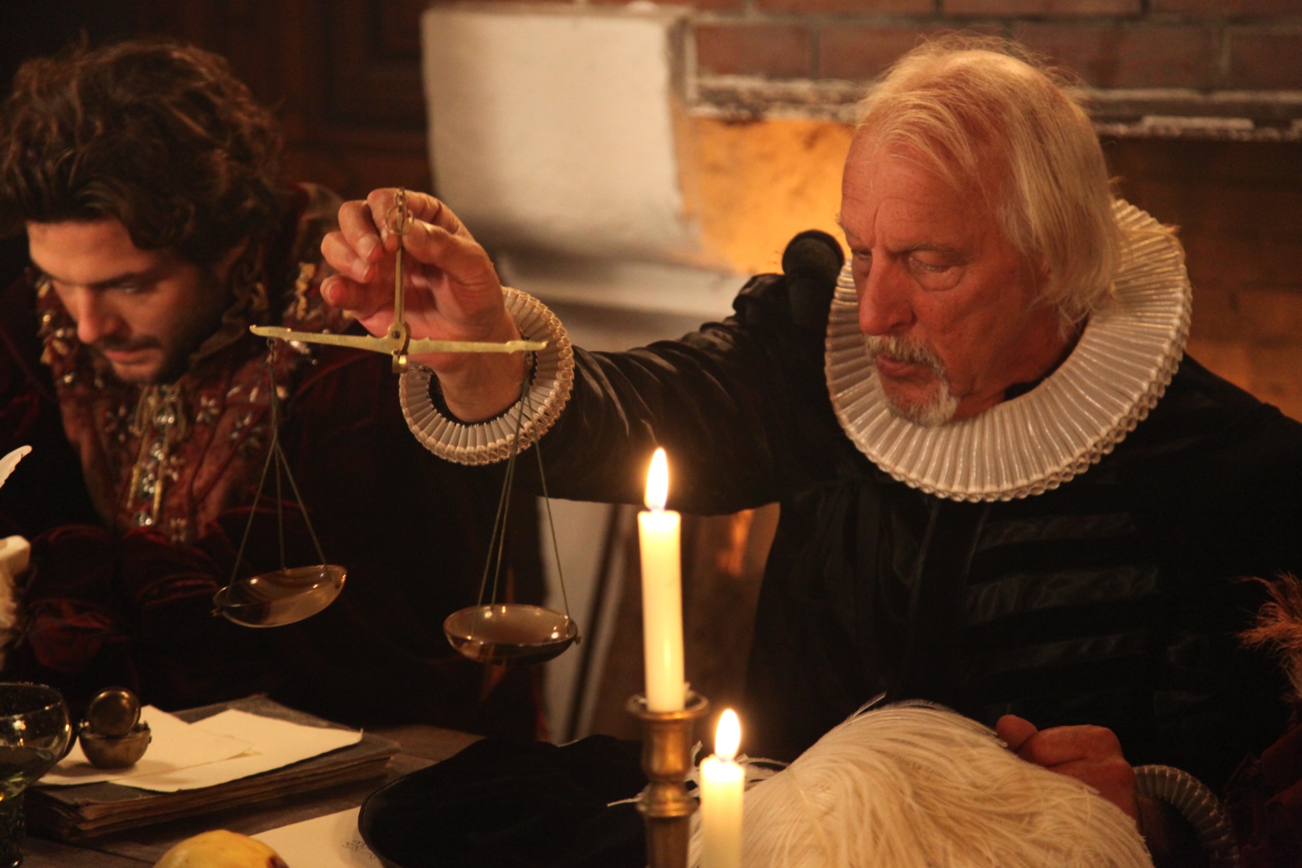 An older man with white hair and a ruffled collar carefully balances a scale over a lit candle in a dimly lit room, reminiscent of the Tulip Mania era. A younger man with dark hair sits beside him, focused on writing about economic bubbles. The setting appears historical and atmospheric.