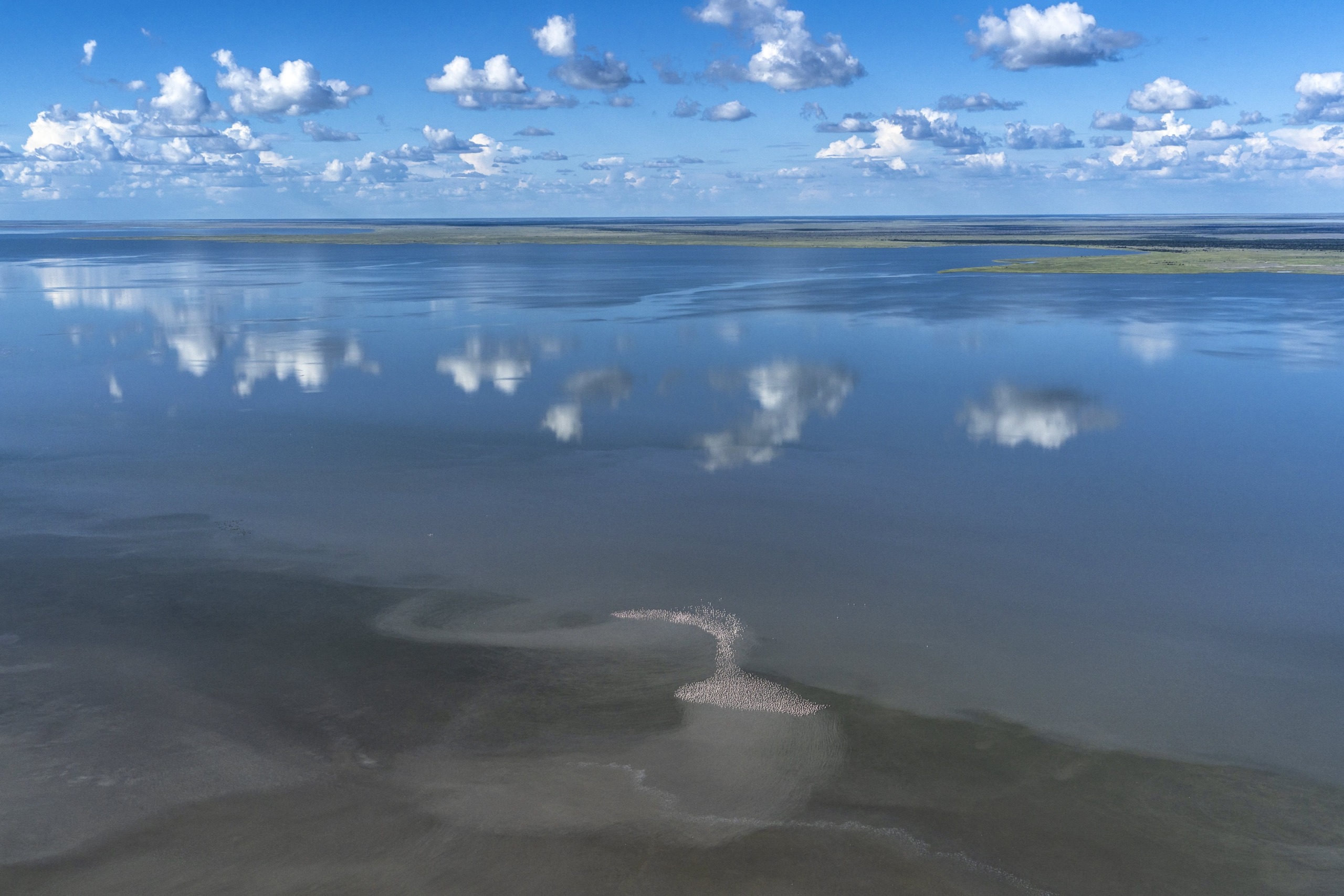 A tranquil aerial view of a vast, high-altitude lake reflects fluffy clouds in the sky. Patches of land are visible on the horizon, and a small flock of birds forms a cluster near the center of the image, creating a sense of serenity and openness.