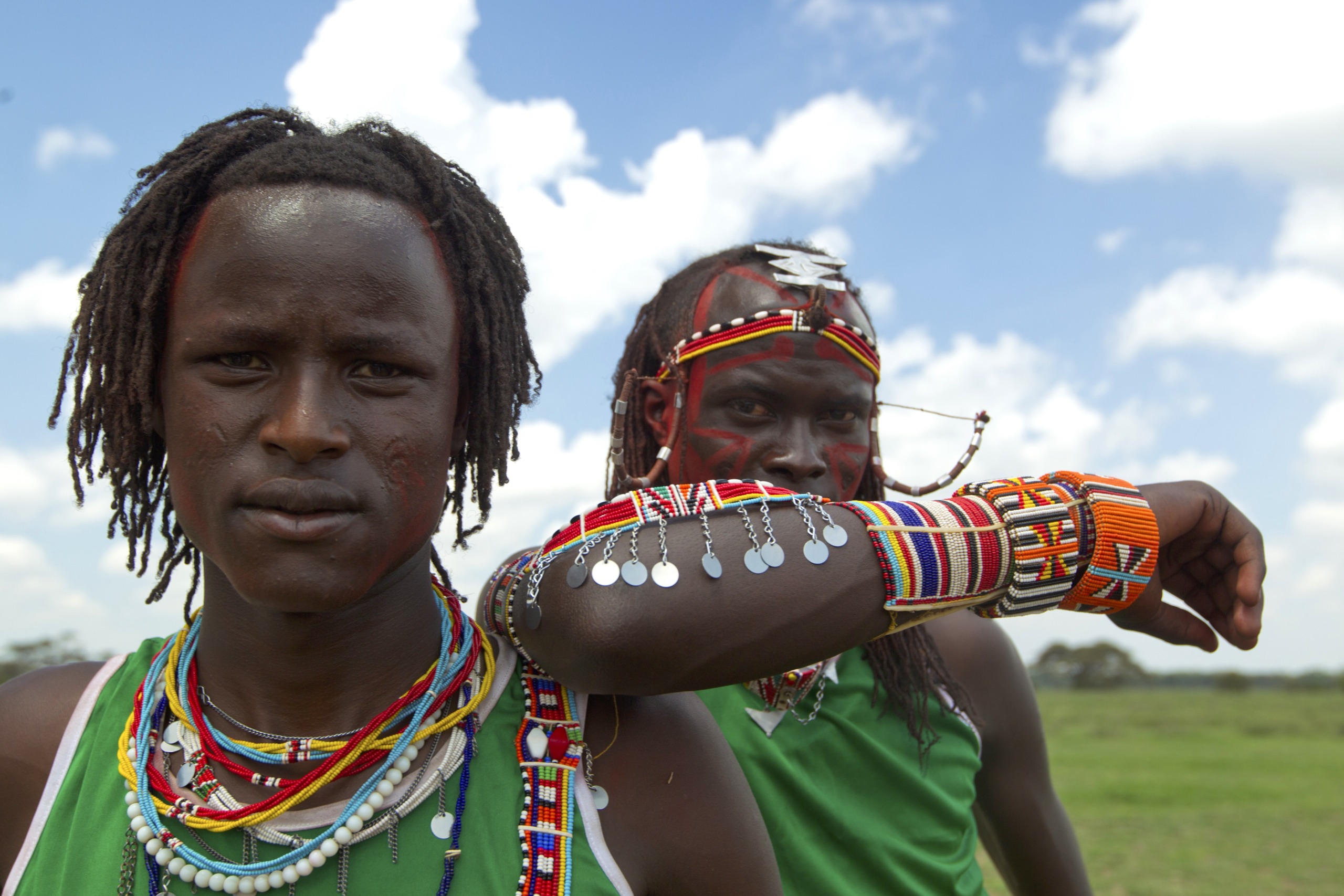 Two individuals in colorful traditional attire and beaded jewelry stand outdoors against a blue sky with scattered clouds, embodying the pride of their tribe. One looks directly at the camera, while the other gazes to the side, with a grassy landscape in the background.